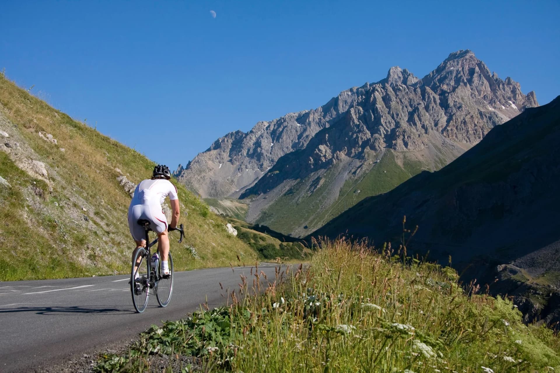 Cyclist in white ascending a mountain road with rocky peaks under a clear blue sky.