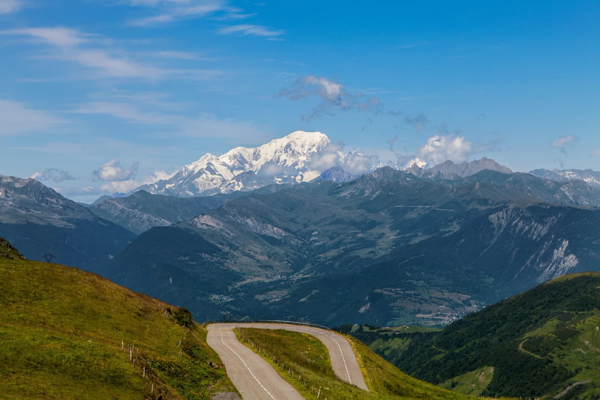 Winding mountain road ascending past green slopes toward snow-capped peaks, likely Col de la Madeleine.