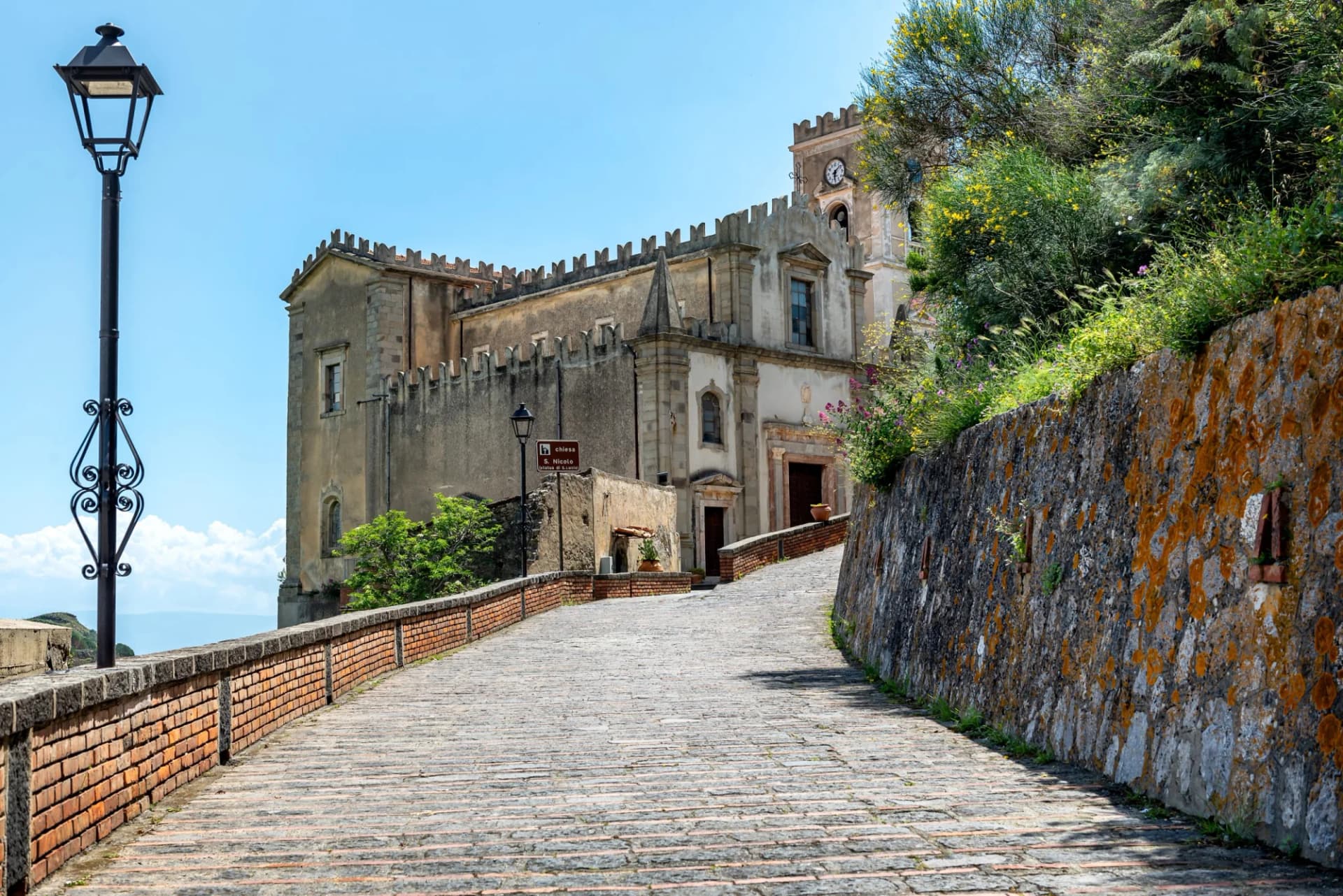 Cobblestone path leading uphill past stone church with crenelated roof and clock tower.