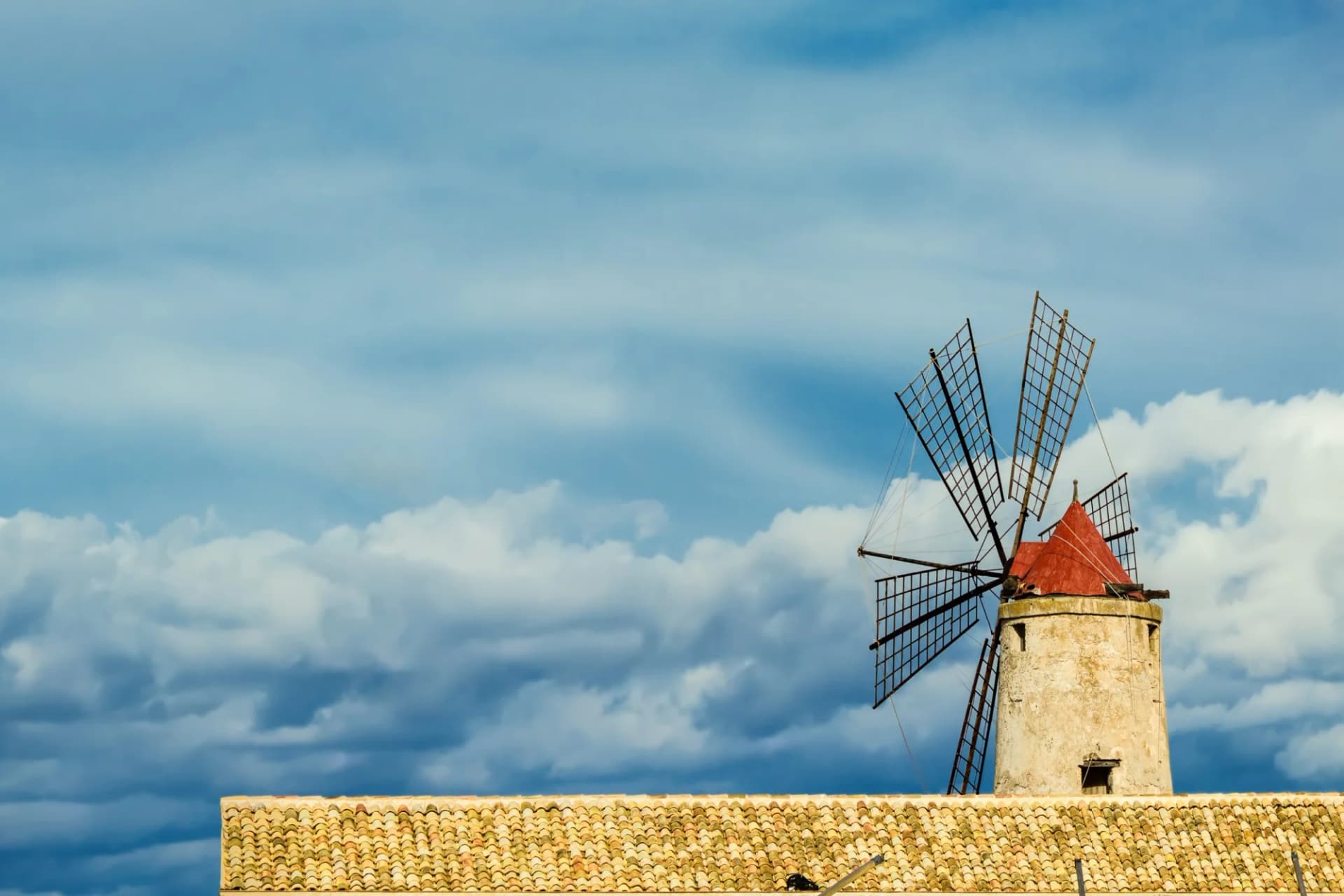 Stone windmill with red cap and lattice sails above a tiled roof against a dramatic cloudy sky.