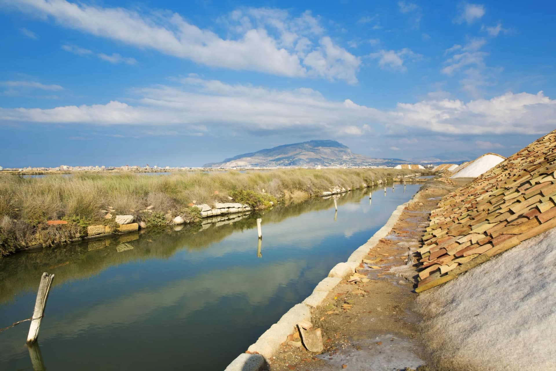 Saltworks canal with salt piles, terracotta tiles, and mountain in background, Trapani, Sicily.
