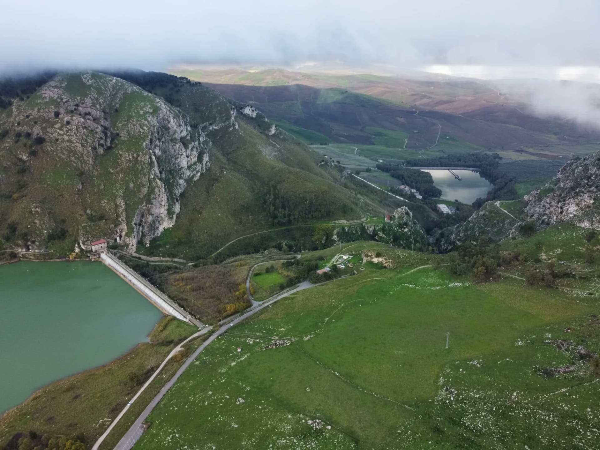 Aerial view of green mountains, a dam, and a green reservoir at Lago Piana degli Albanesi.