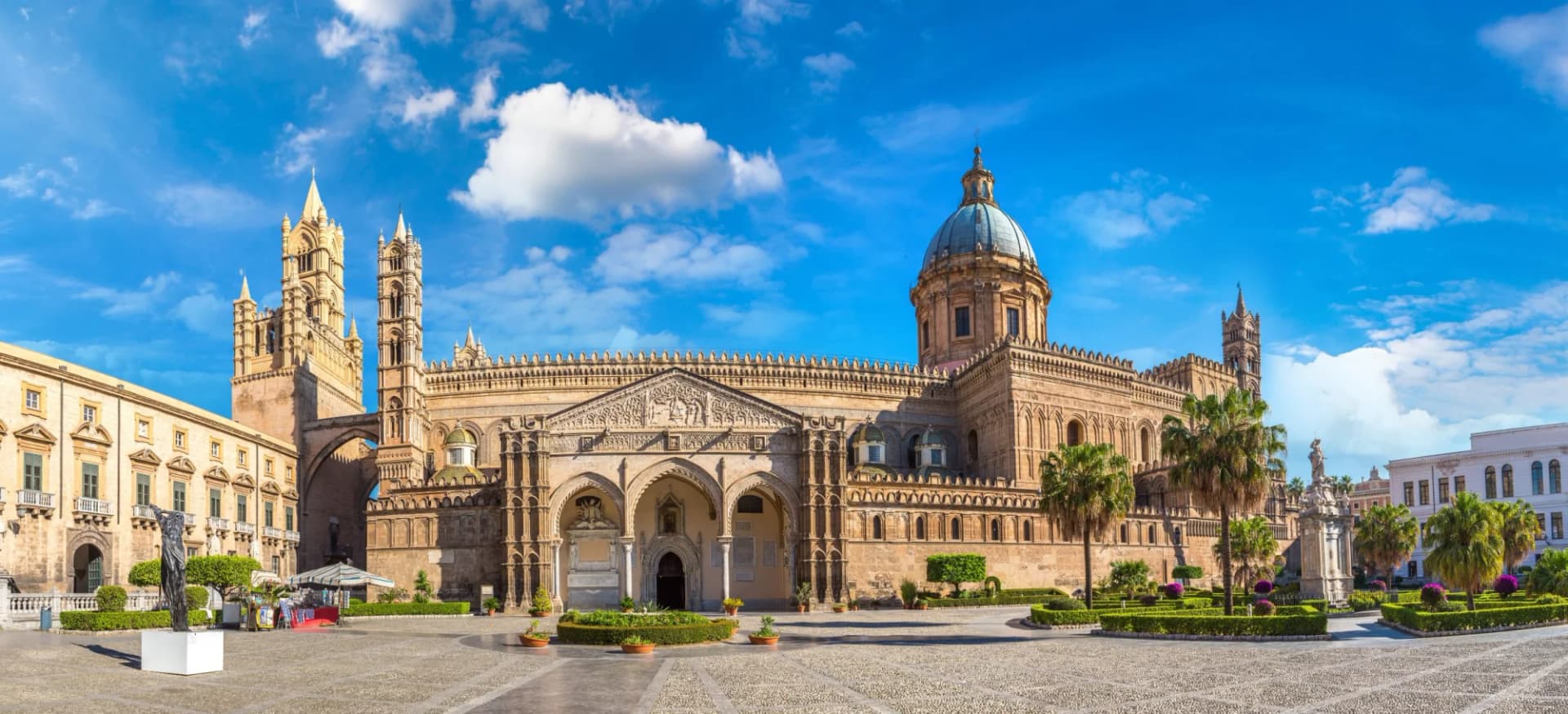 Palermo Cathedral facade with towers and dome under a bright blue sky with clouds.