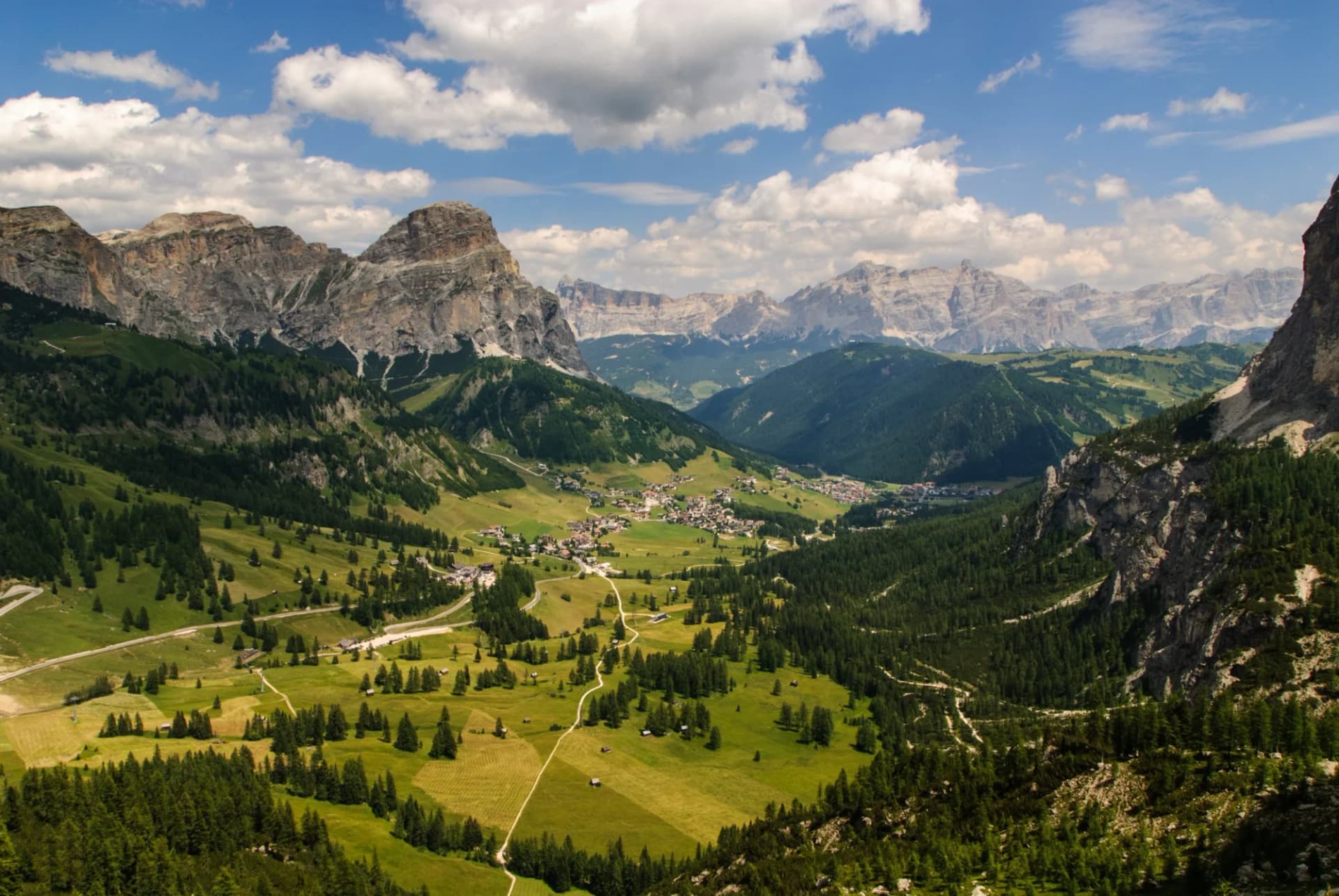 Alpine valley with green meadows, pine forests, and a village nestled below rocky peaks in Val di Fassa.