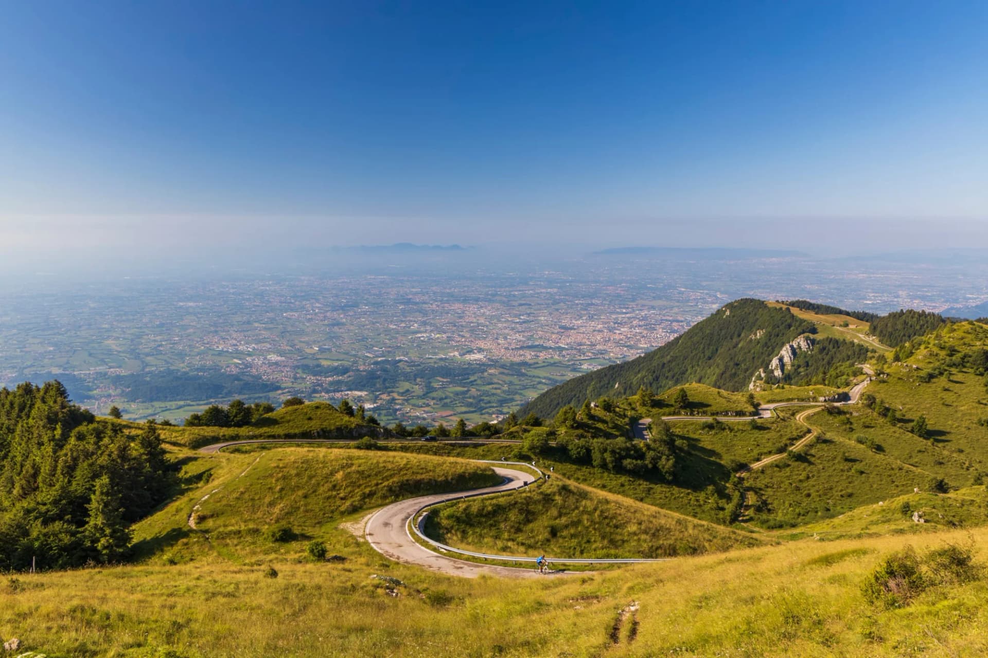 Winding mountain road with a cyclist overlooking a vast, hazy plain and city below, Monte Grappa.