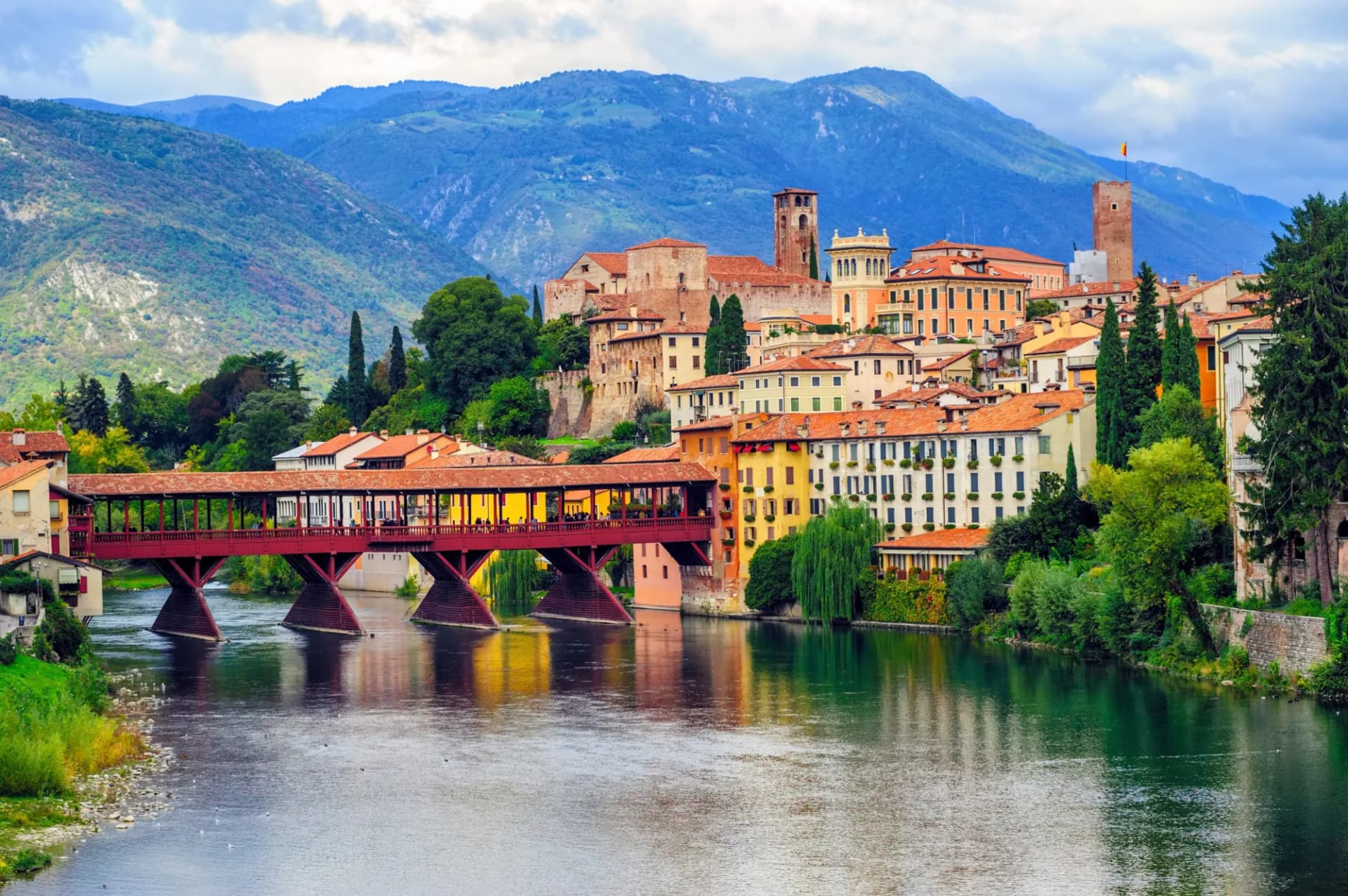 Covered wooden bridge over river in Bassano del Grappa with colorful buildings and mountains.