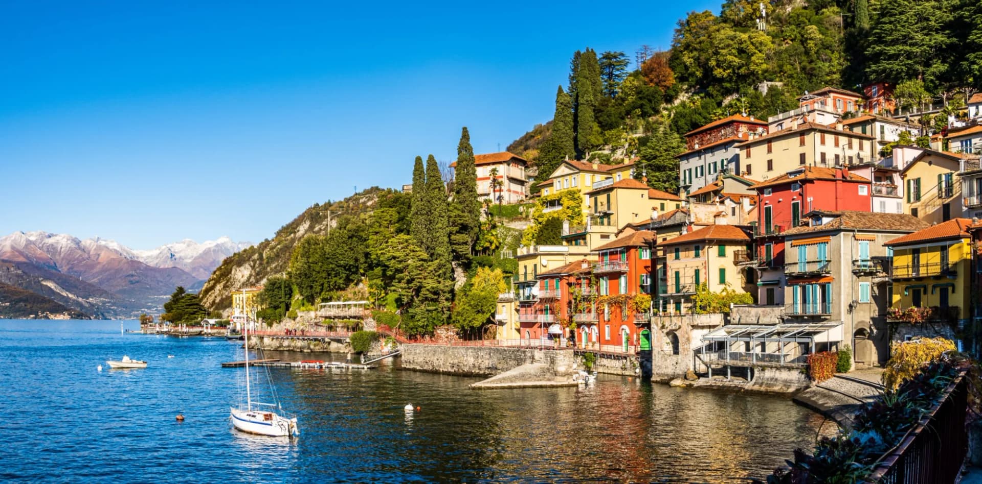 Colorful buildings on steep hillside above Lake Como with snow-capped mountains in distance.