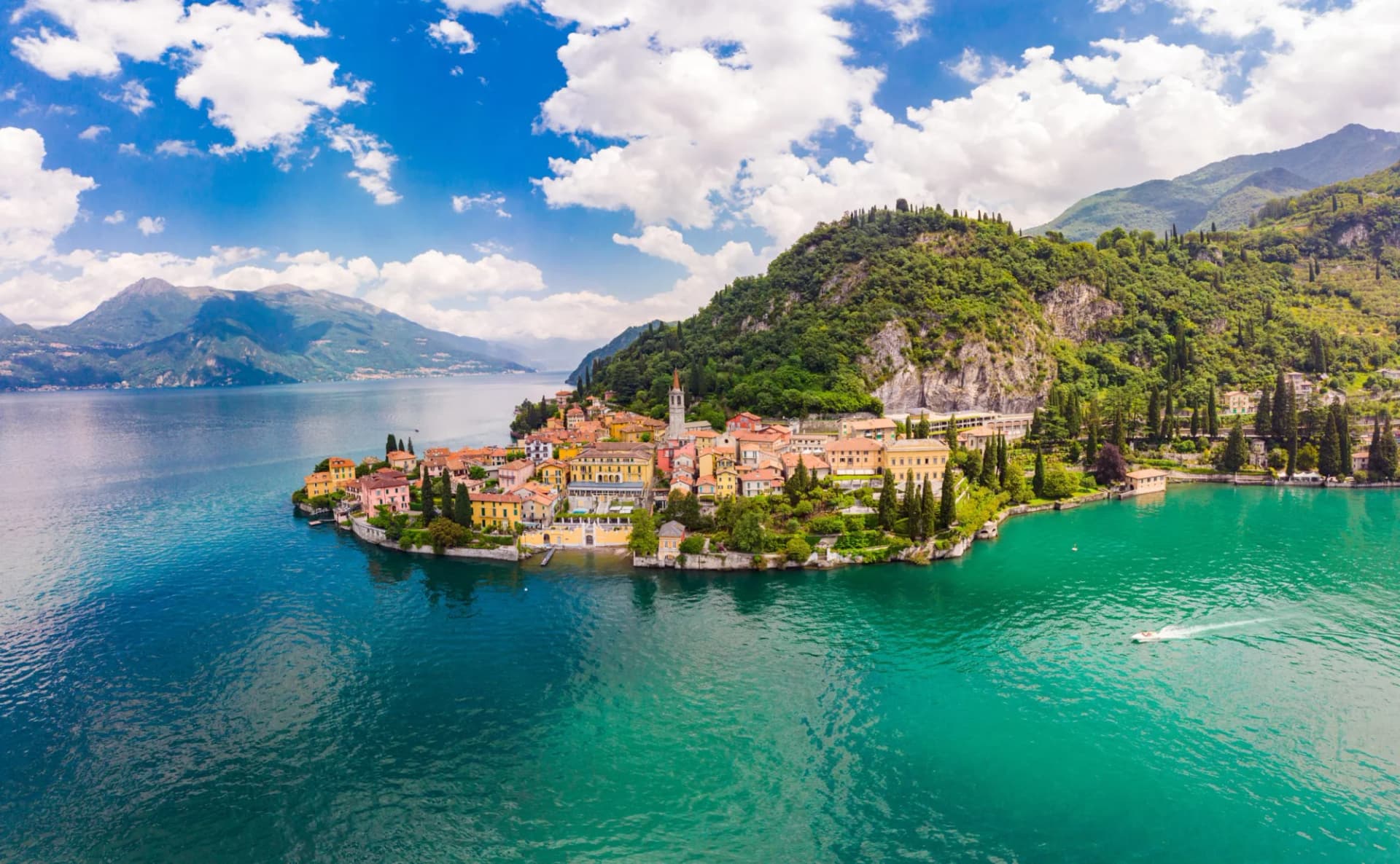 Colorful village on peninsula jutting into Lake Como with mountains and blue sky.