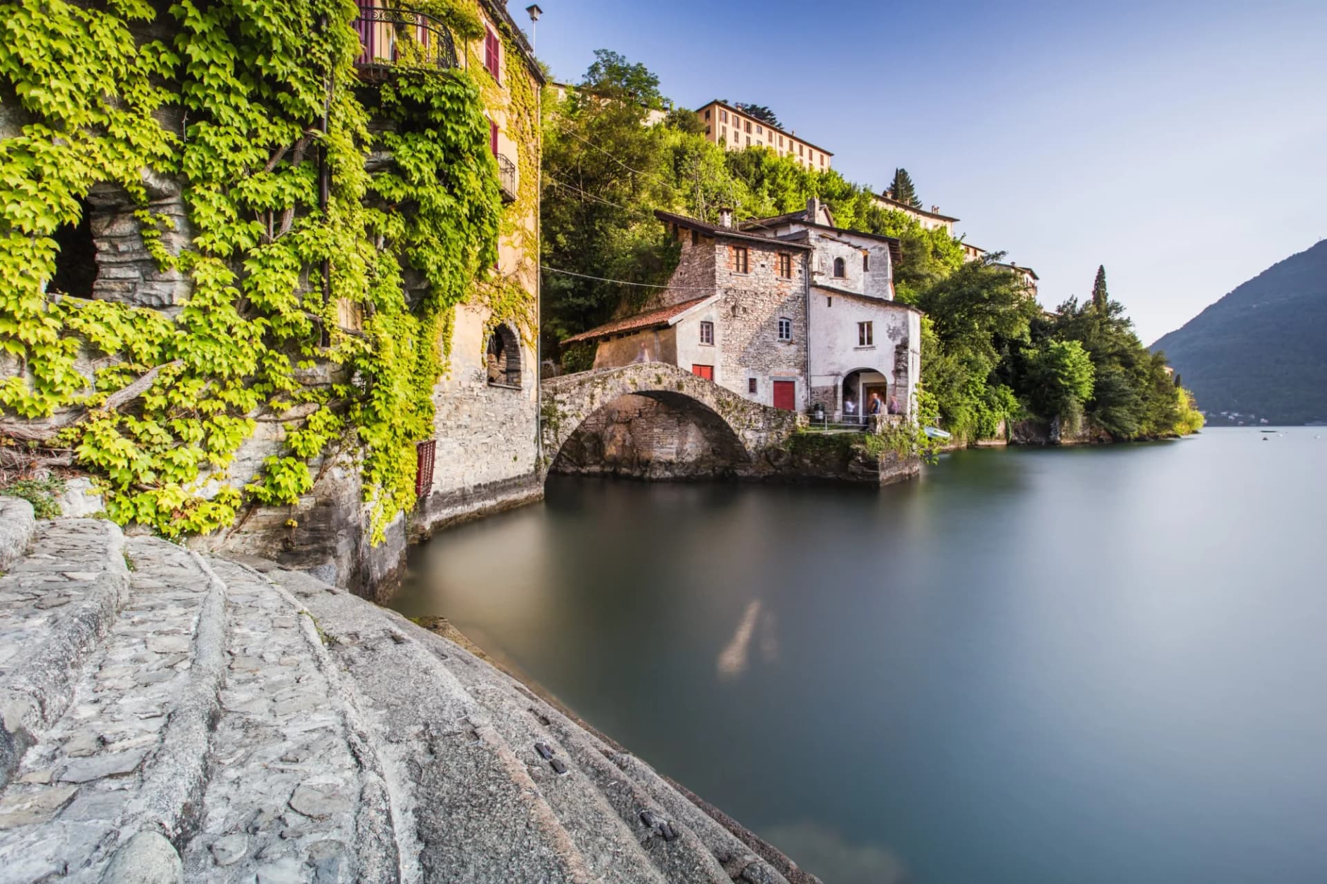 Stone steps along ivy-covered buildings and an arched bridge over calm water at Lake Como.