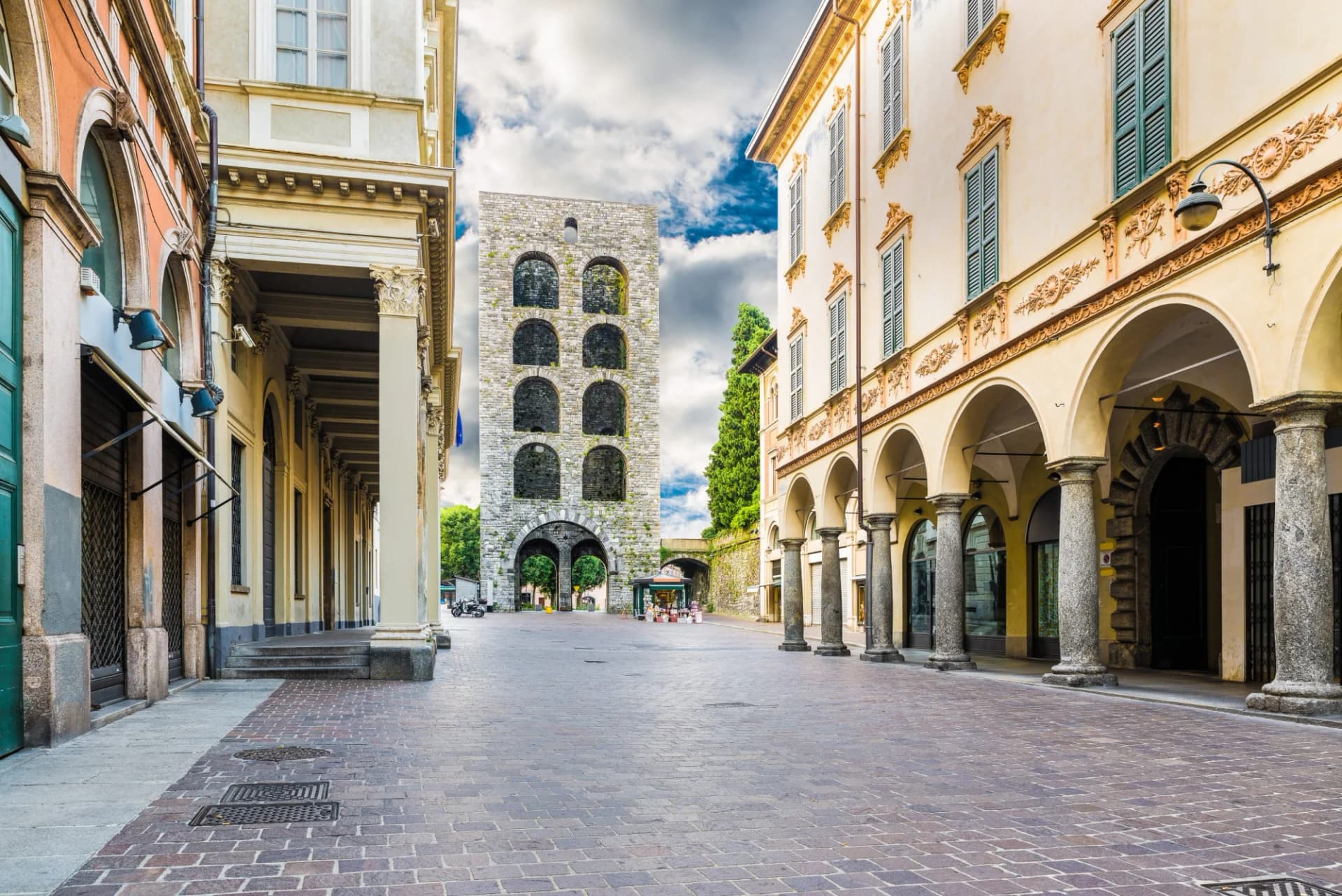 Cobblestone square with historic stone tower and arcaded buildings under cloudy sky.