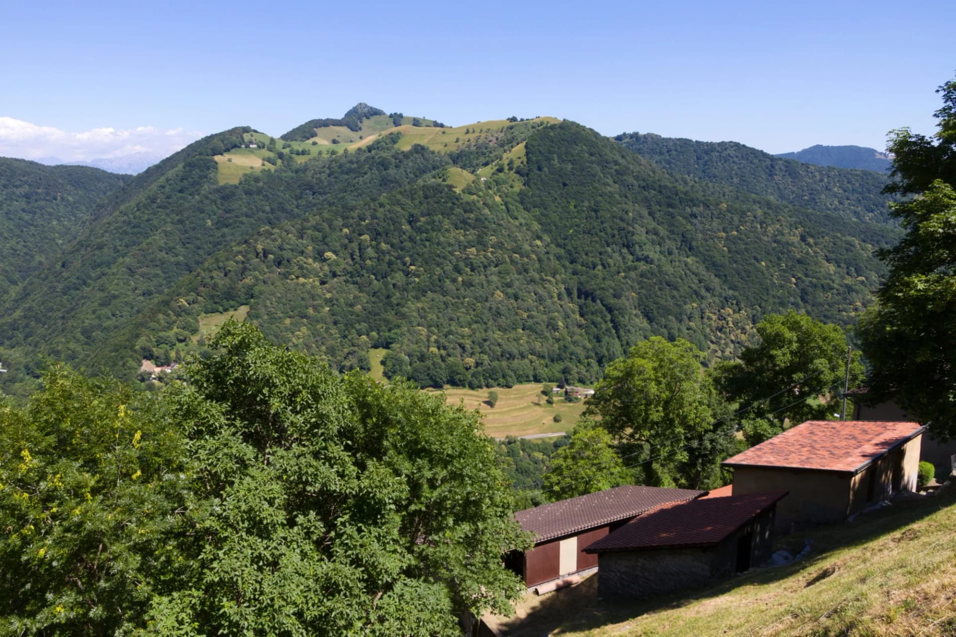 Small buildings with terracotta roofs on grassy slope overlooking lush green mountains under clear blue sky.