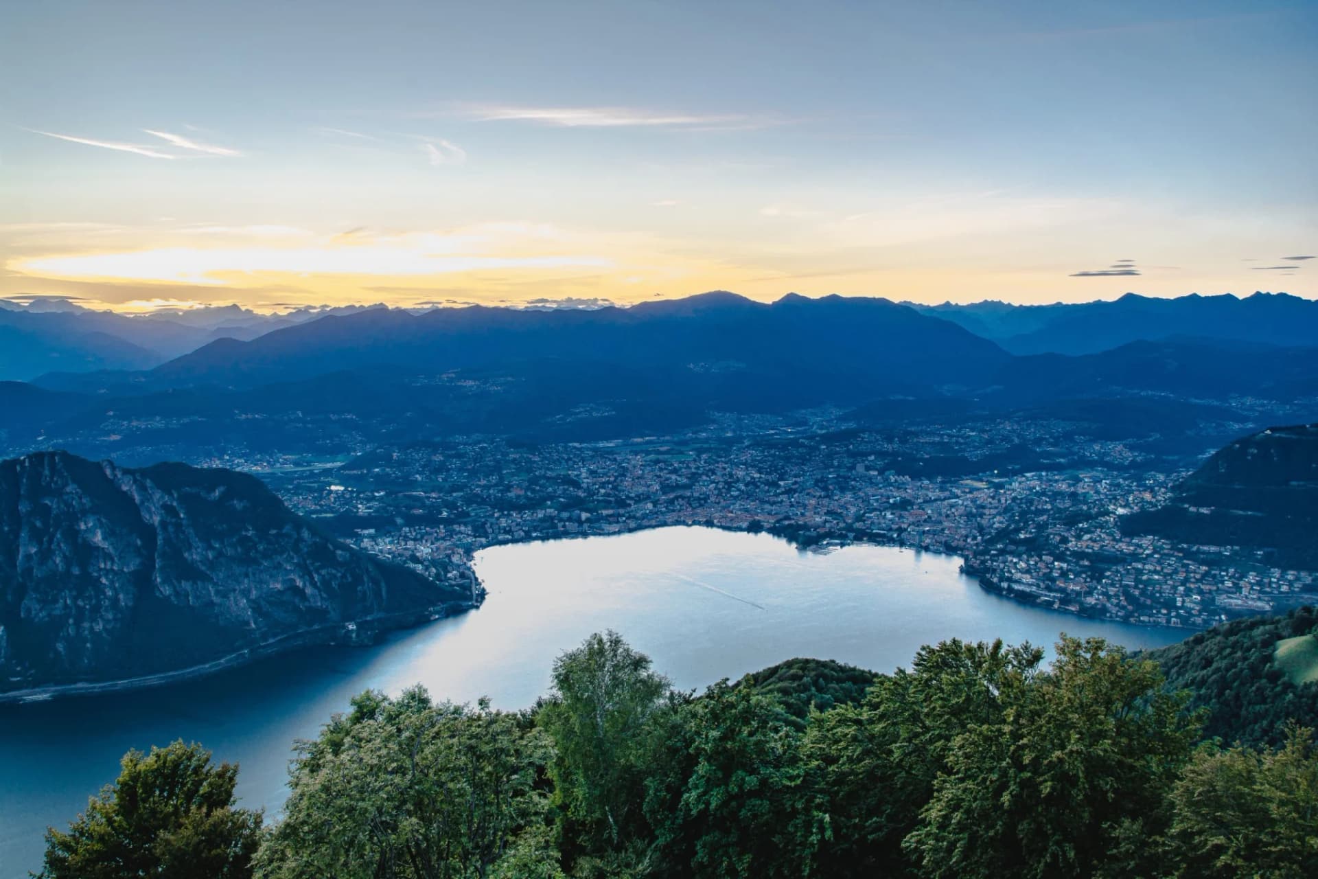 Panoramic view of a city nestled around a lake, surrounded by dark mountains at sunset.