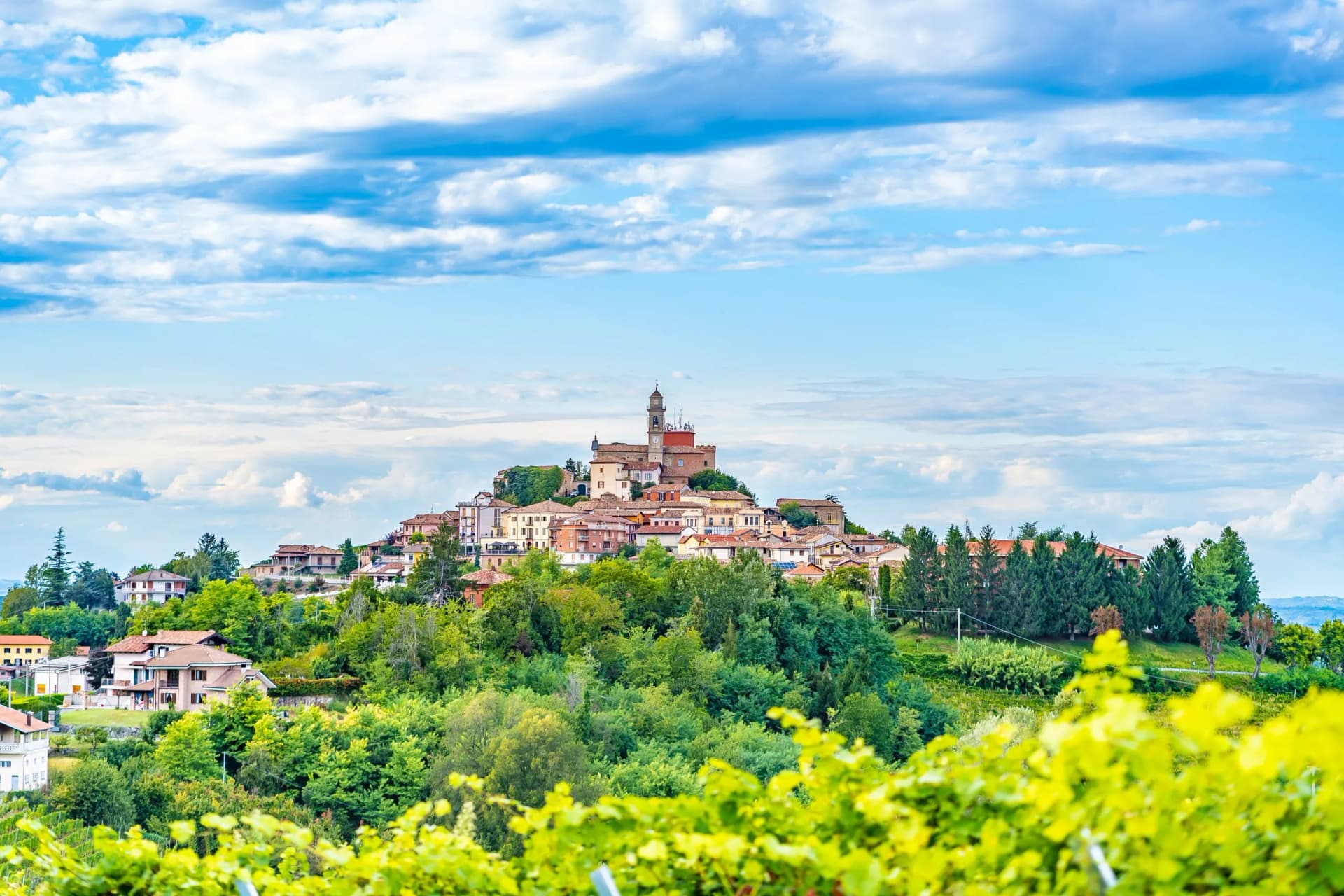 Hilltop town with church tower overlooking lush green vineyards under a blue, cloudy sky.