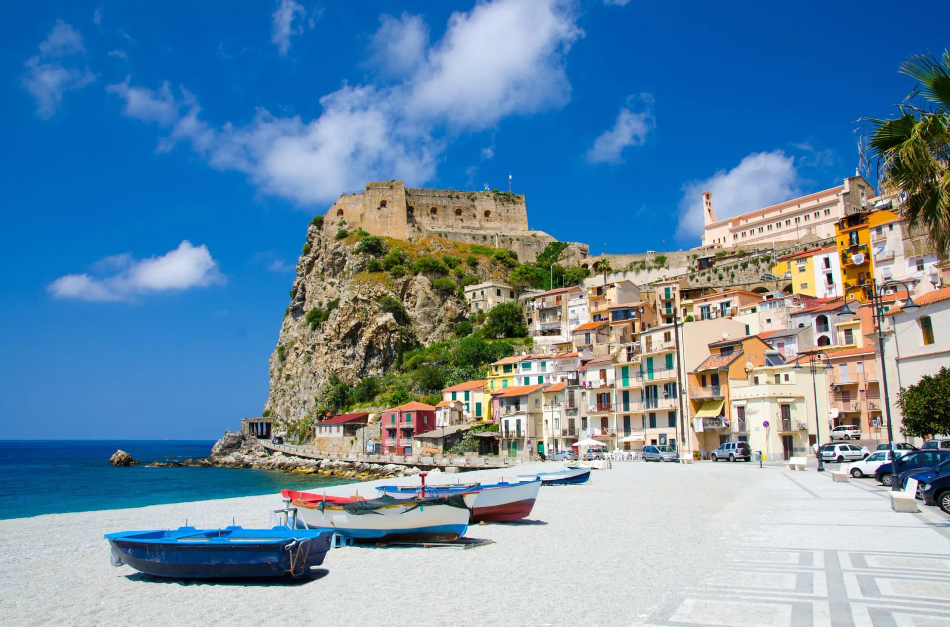 Fishing boats on a white pebble beach below a cliffside town and fortress in Sicily.