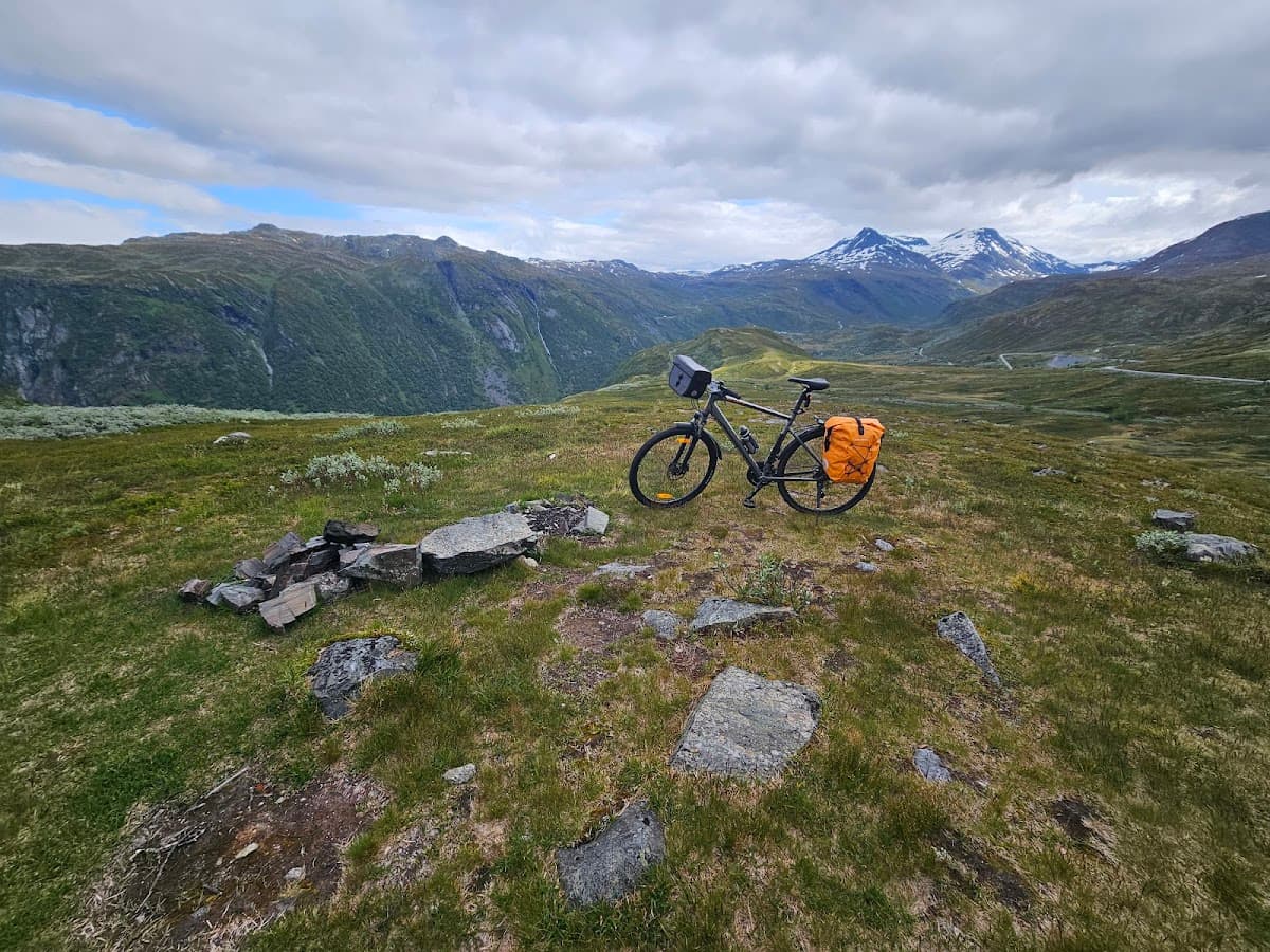 Bicycle touring with orange panniers on grassy mountain overlook with snow-capped peaks.