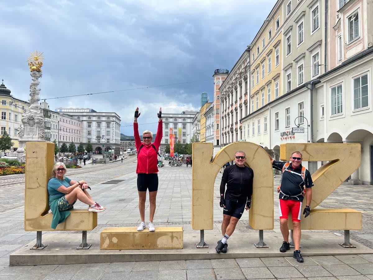 Four cyclists pose with large golden "LINZ" letters in a European city square.