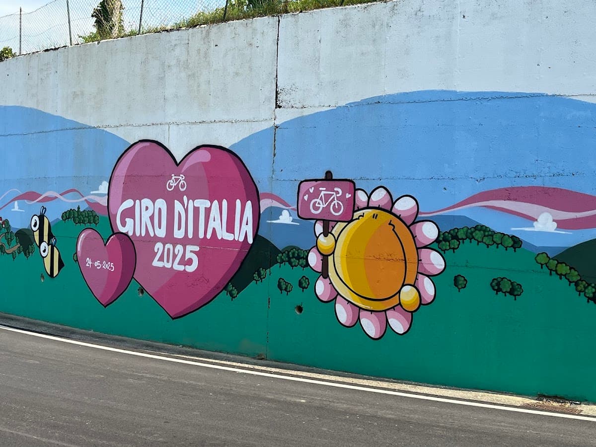 Giro d'Italia 2025 mural with hearts, bees, and a sun character by a roadside.
