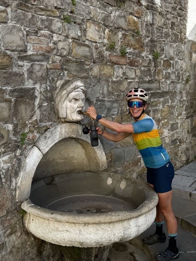 Cyclist filling water bottle at historic stone fountain set into a rough stone wall