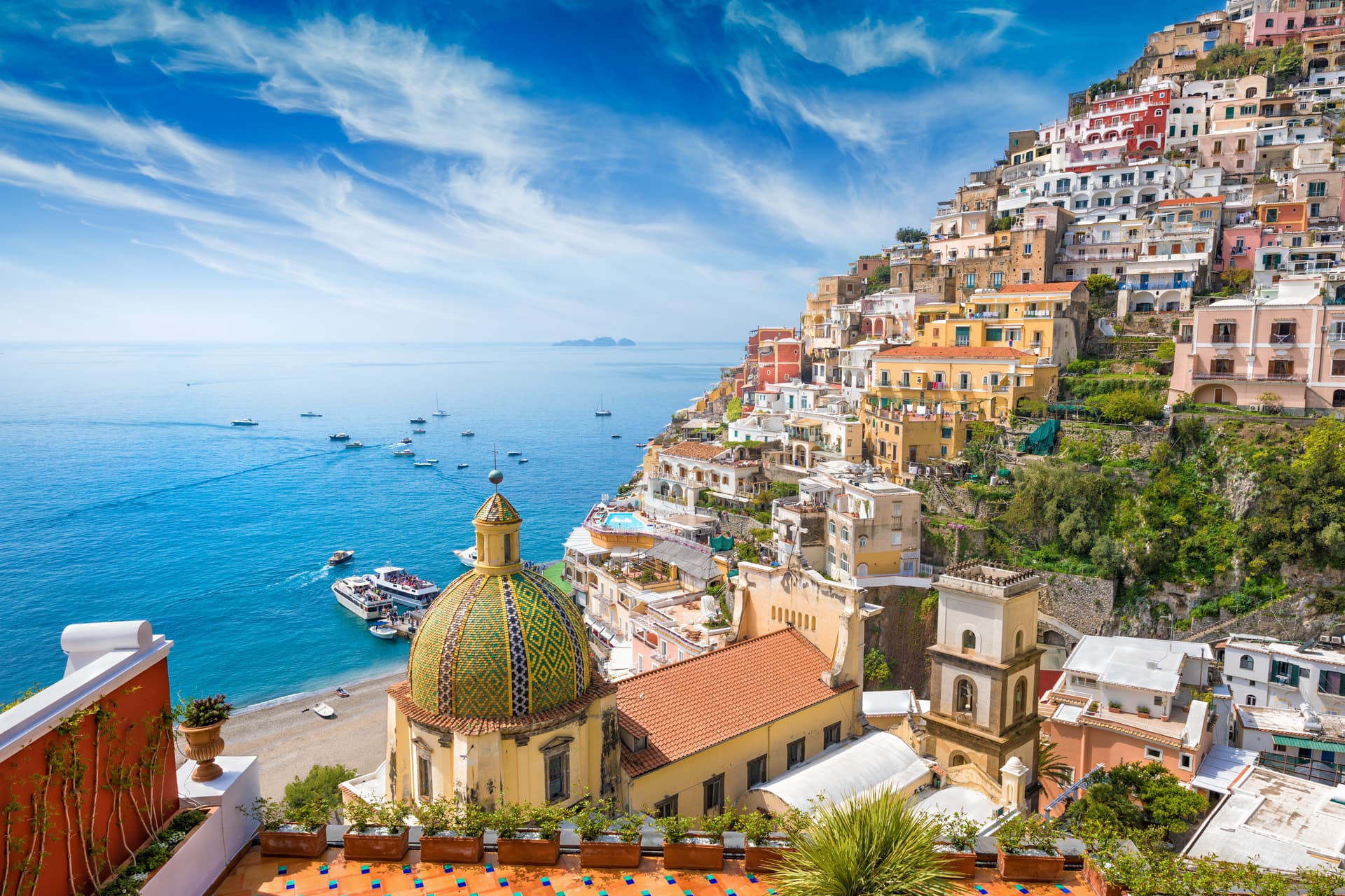 Positano cathedral dome overlooking colorful cliffside buildings and boats on the blue sea.