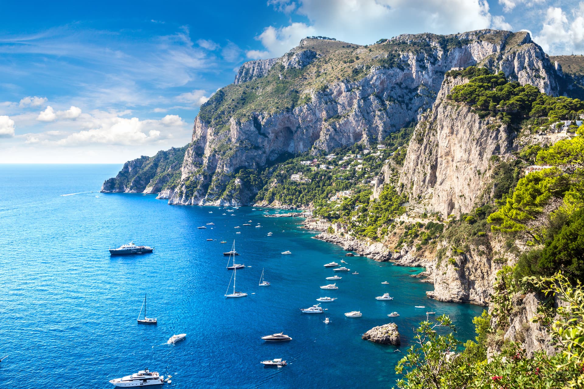 Boats anchored in turquoise water below steep, green cliffs of Capri under a blue sky.