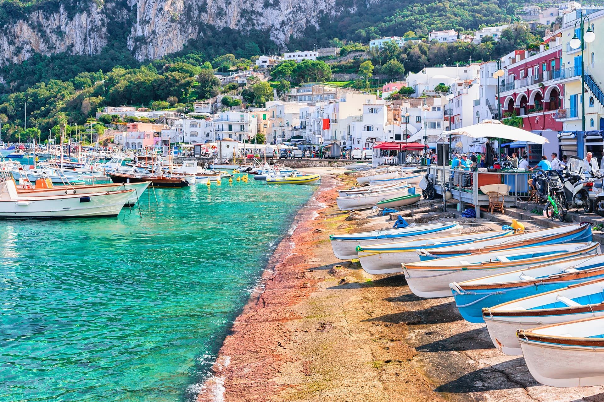 Small boats pulled ashore next to turquoise water in Capri port with white buildings climbing a cliff.