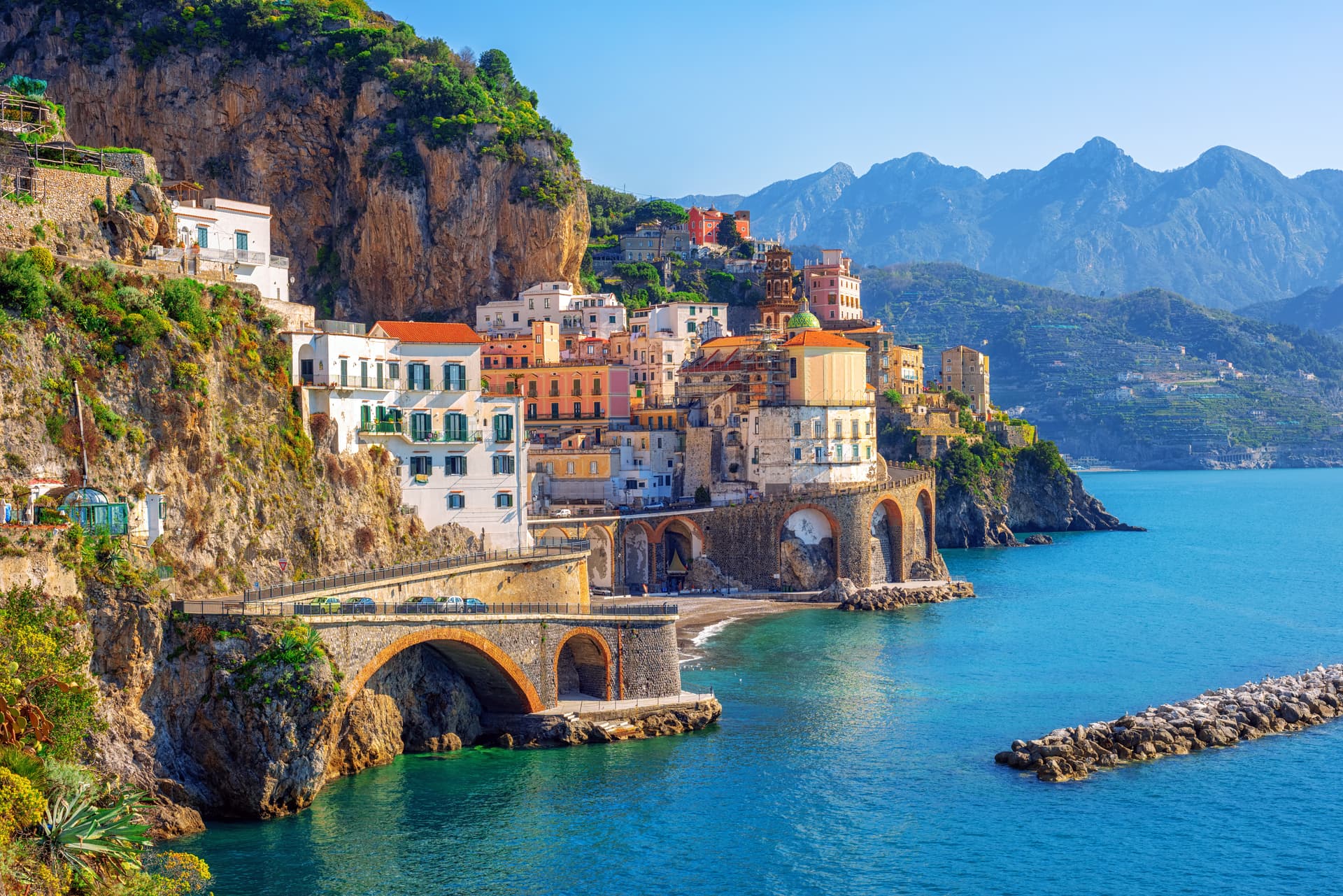 Colorful buildings clinging to cliffs above turquoise water in Atrani, Amalfi Coast, with mountains in background.