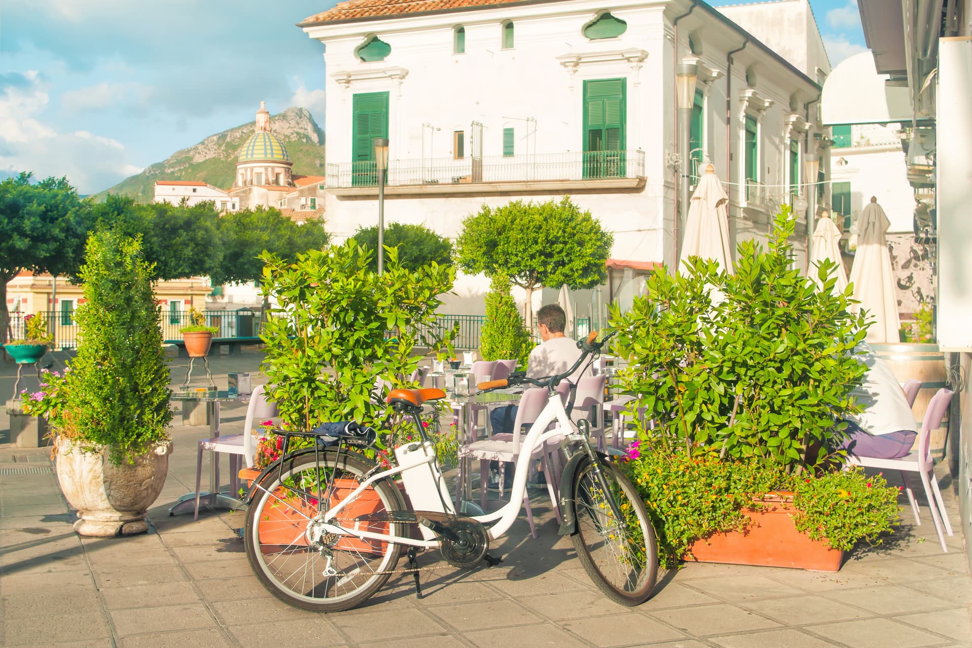 White bicycle parked near outdoor cafe seating with lush greenery and mountain view.