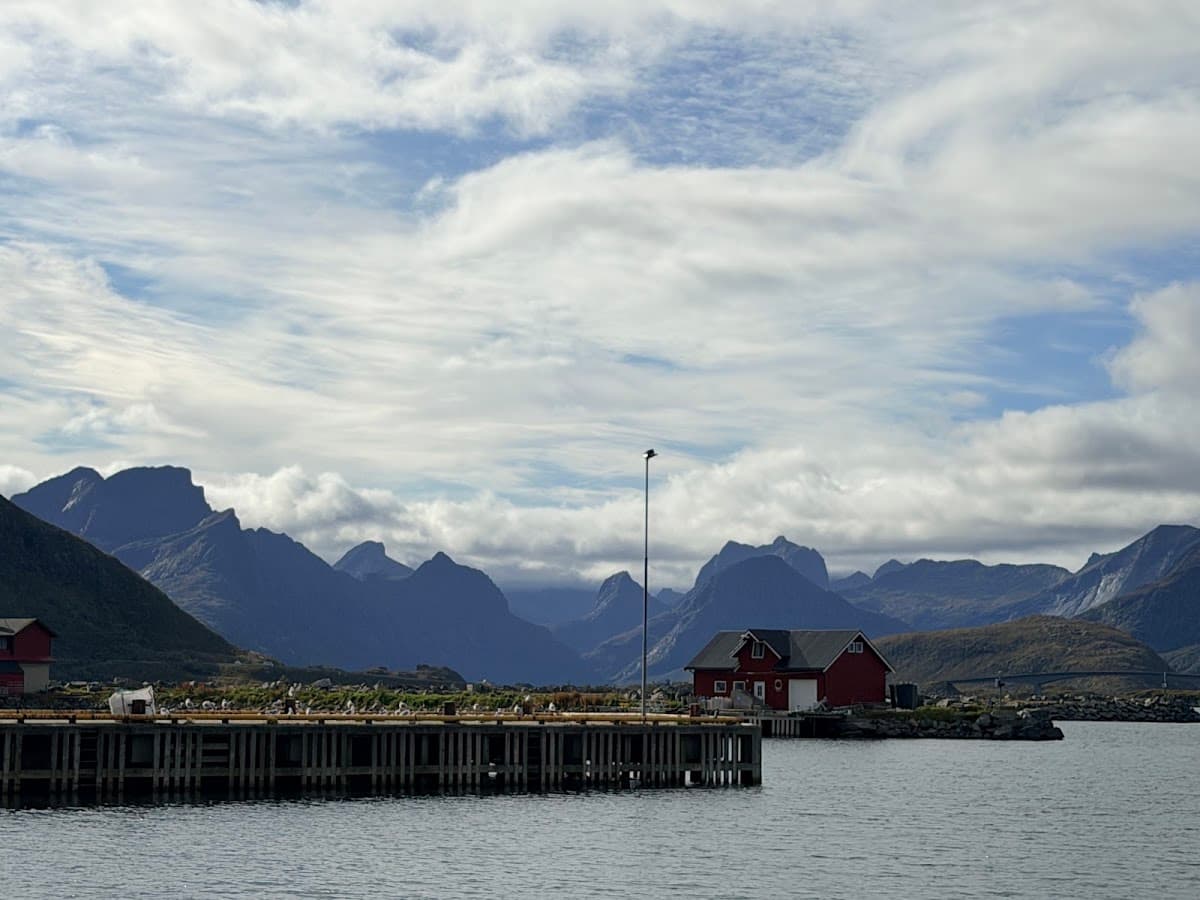 Wooden pier and red waterfront building beneath dramatic mountain peaks and cloudy sky