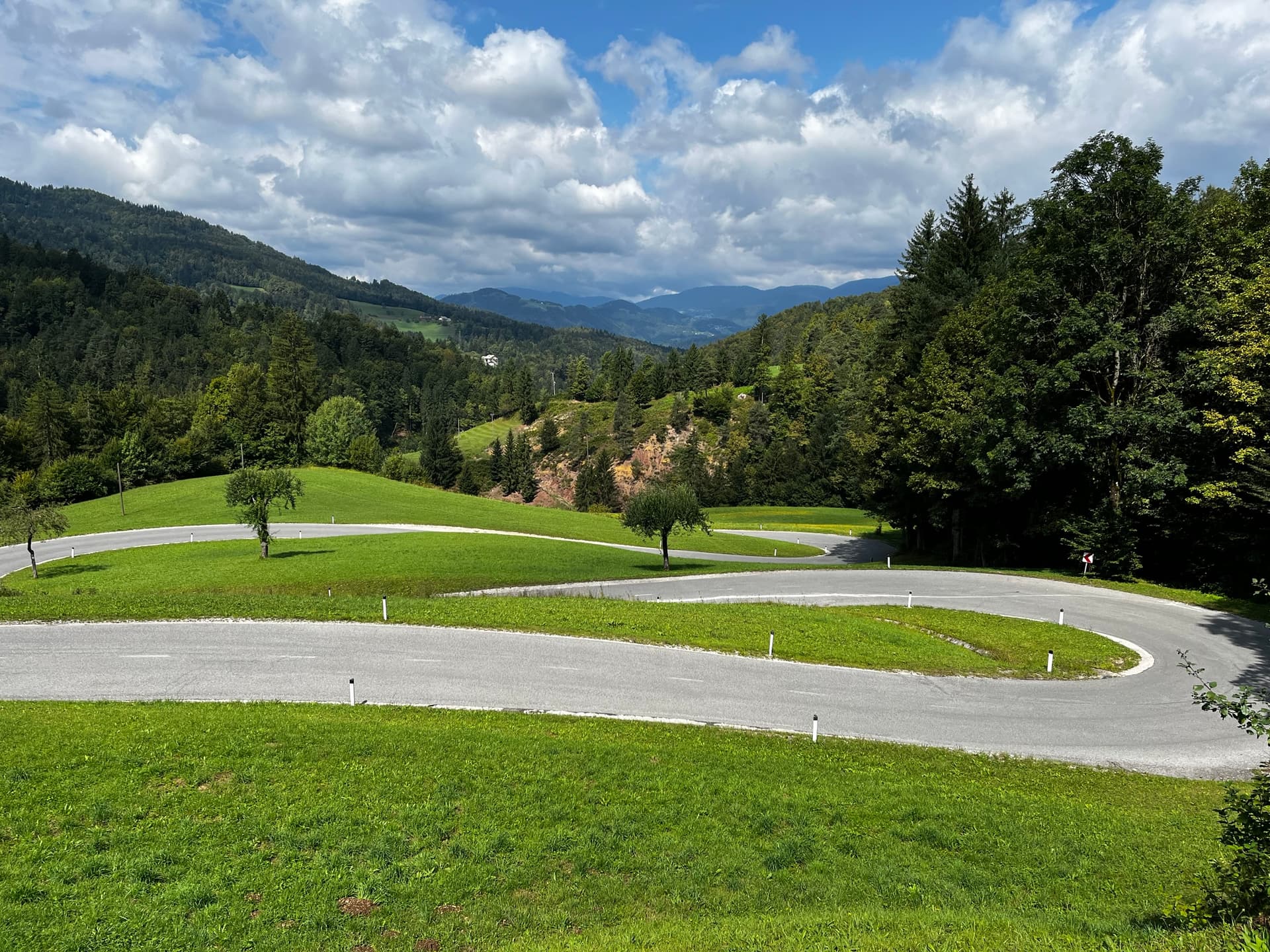 Hairpin road curving through green meadows and forested hills under a cloudy sky