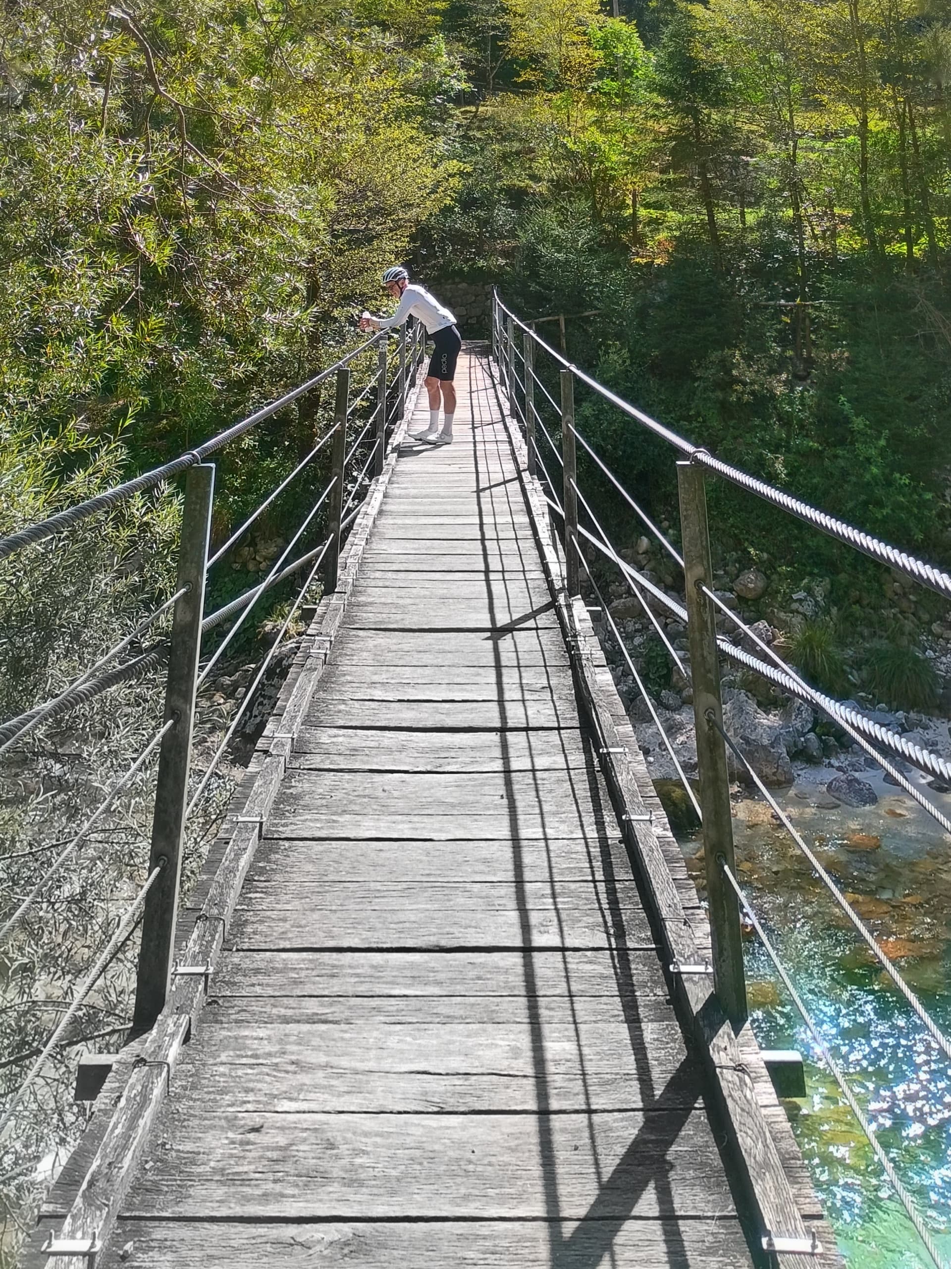 Cyclist standing on a wooden suspension footbridge over a clear river in a forest.