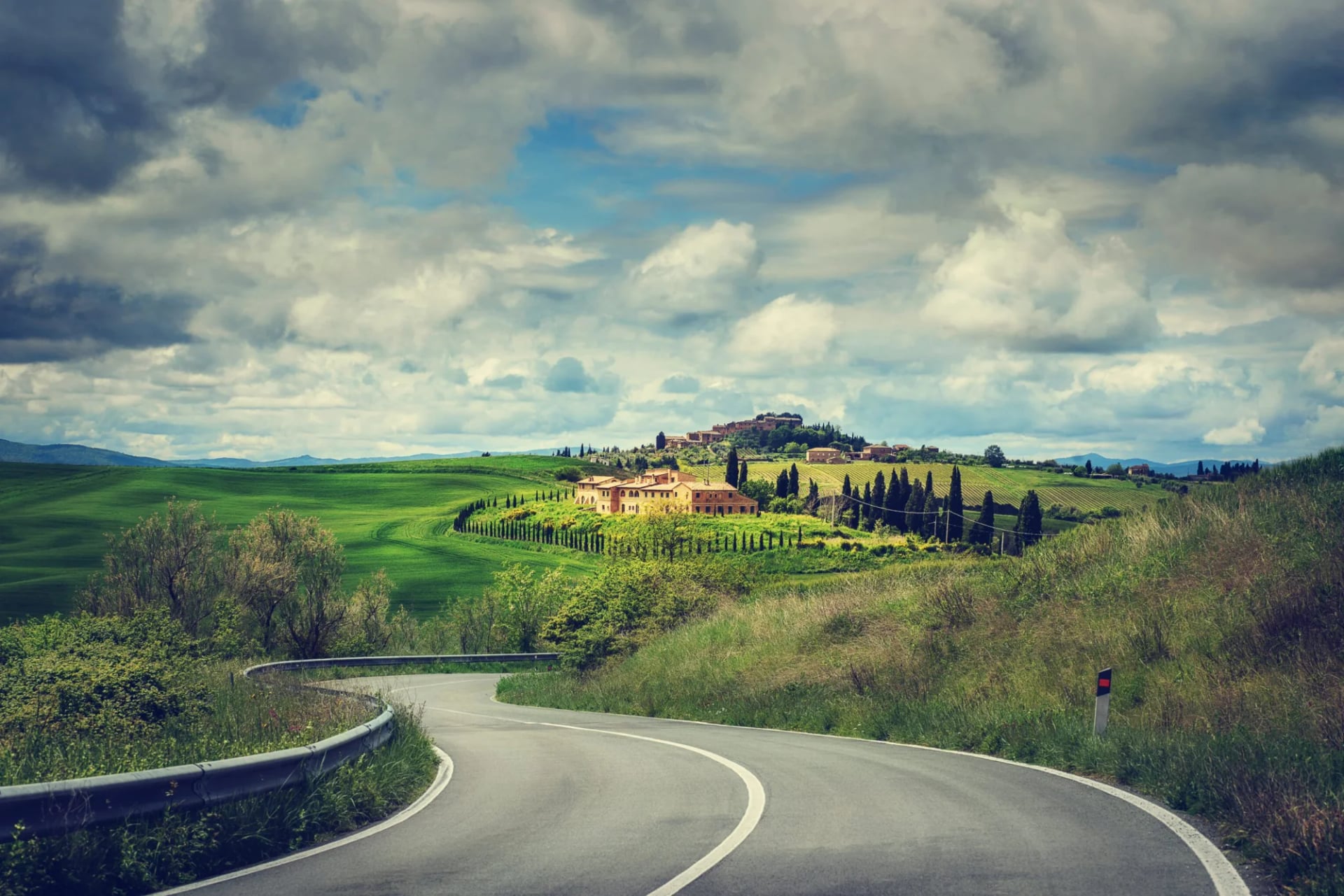 Winding road through green rolling hills toward a Tuscan villa under cloudy skies