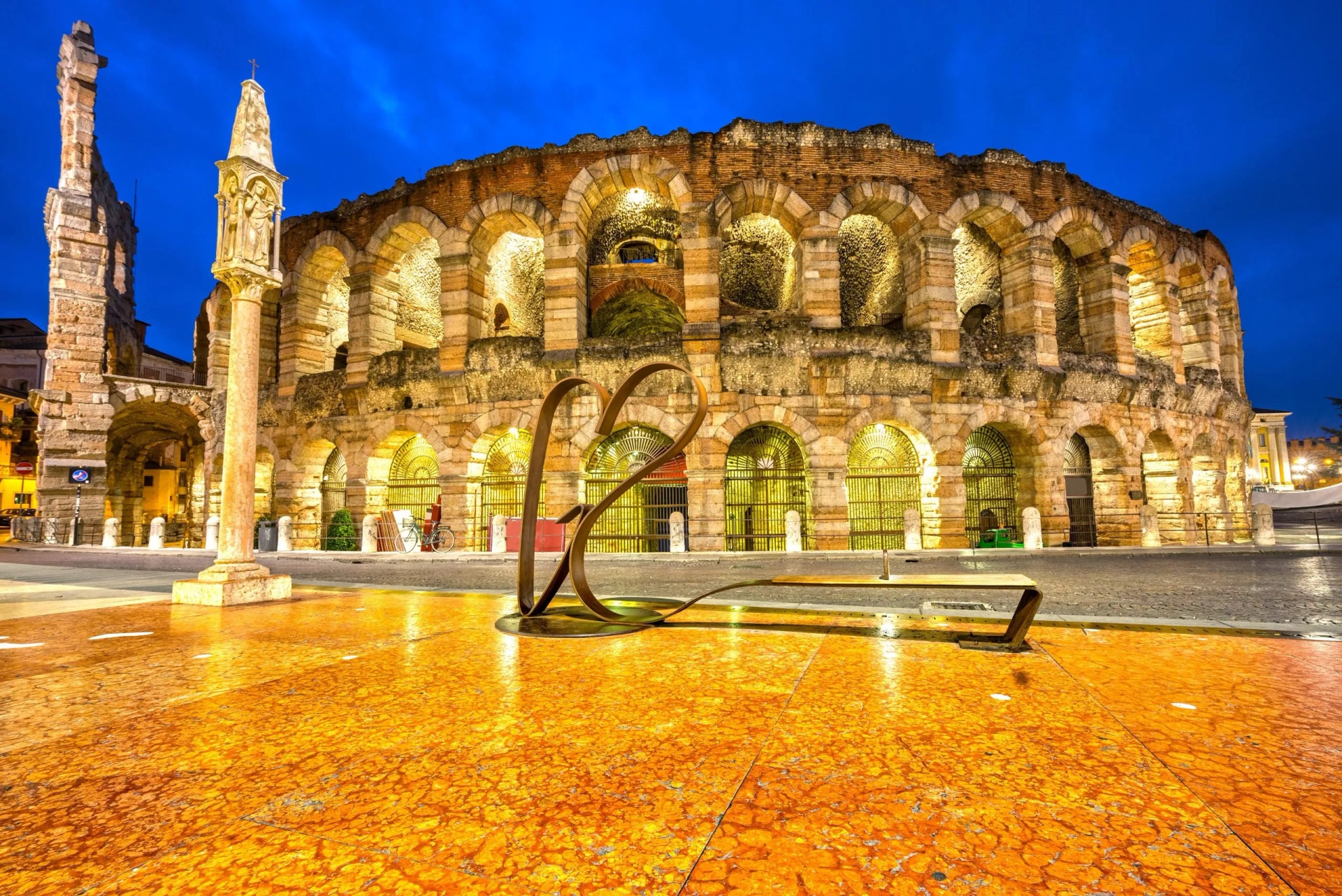 Verona Arena illuminated at night with a heart sculpture in the foreground and blue sky.