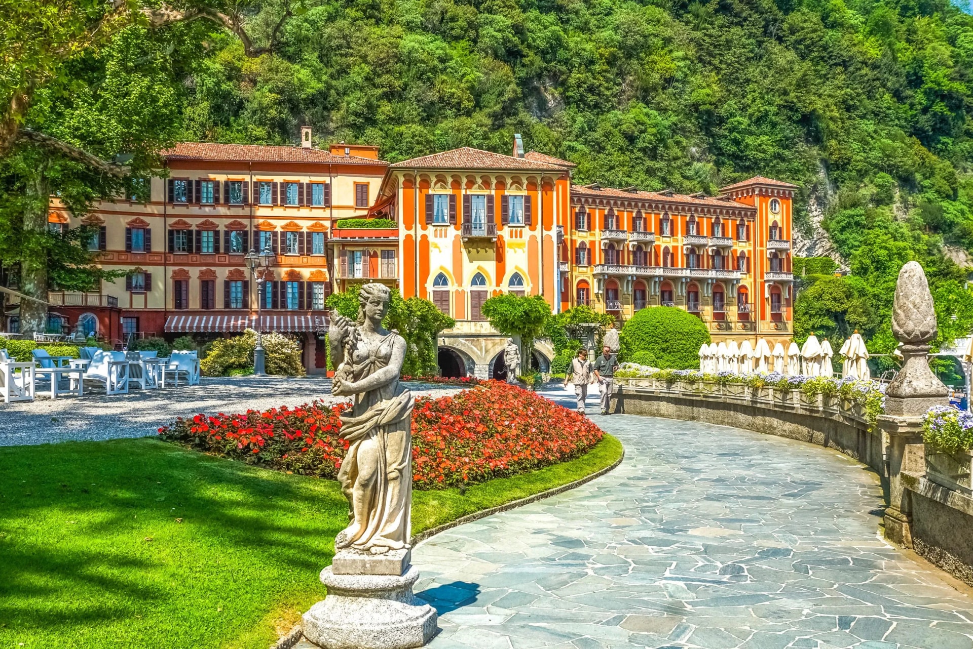 Statue in garden before colorful hotel building against lush green mountain hillside.