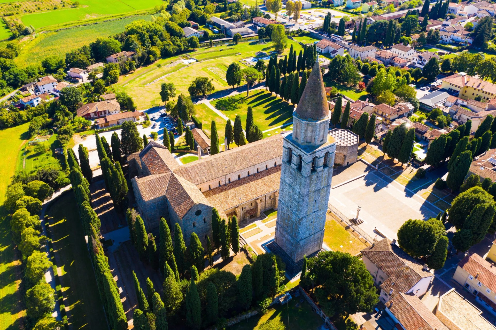 Aerial view of Aquileia center with large stone basilica, tall bell tower, and surrounding green fields.