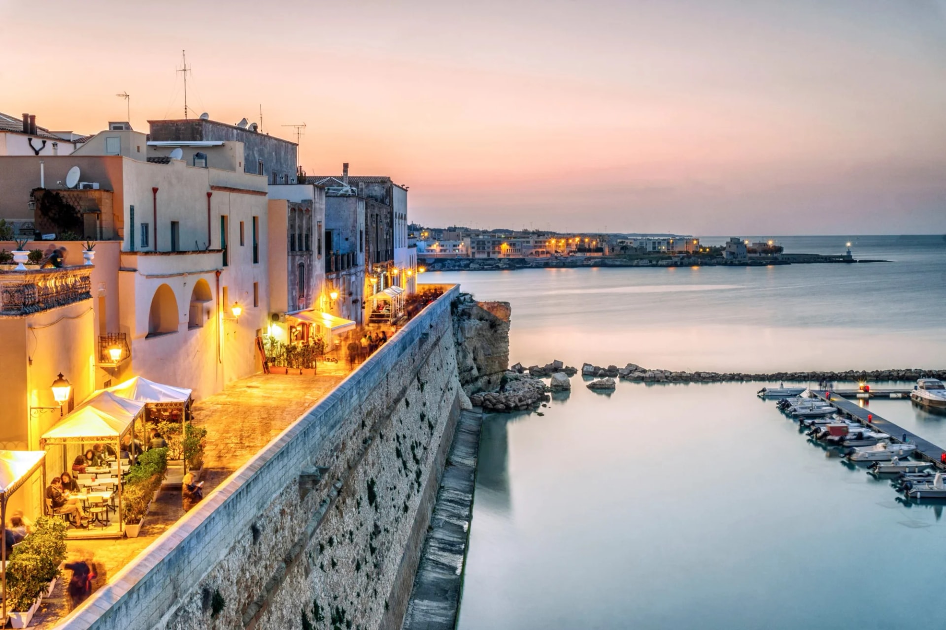 Waterfront dining along a stone wall overlooking a marina at dusk in Otranto, Puglia, Italy.