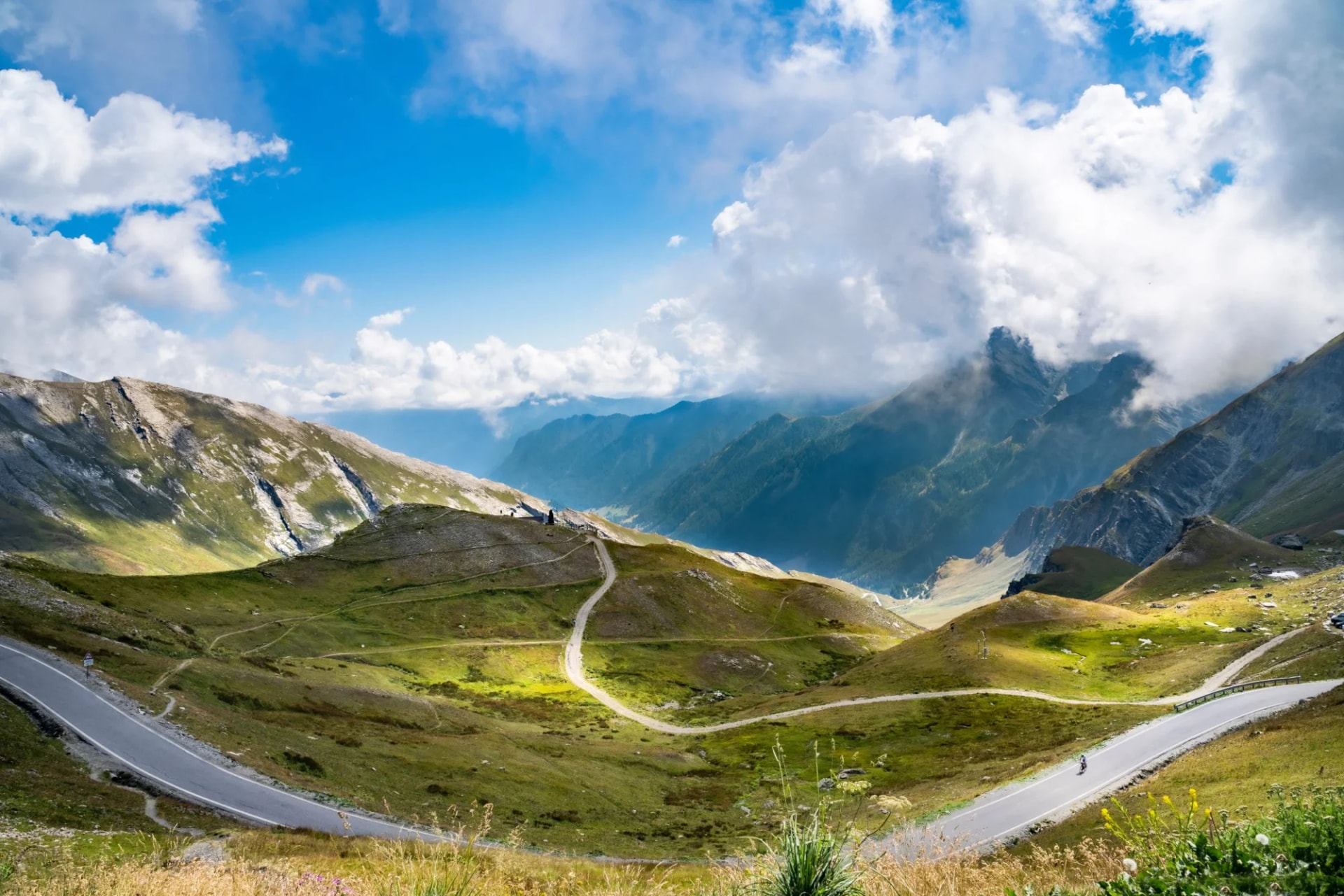 Winding mountain road with switchbacks through green hills under a dramatic blue sky with clouds.