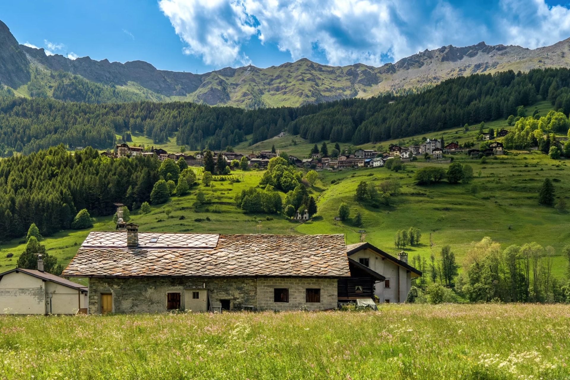 Stone farmhouse in a grassy field with a village and green mountains in Aosta Valley.