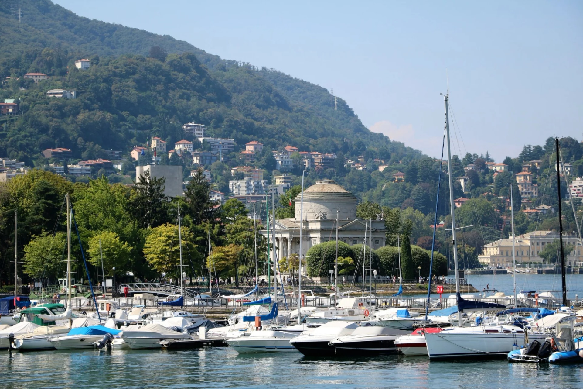 Sailboats docked in a harbor with a domed neoclassical building and lush green mountain backdrop.