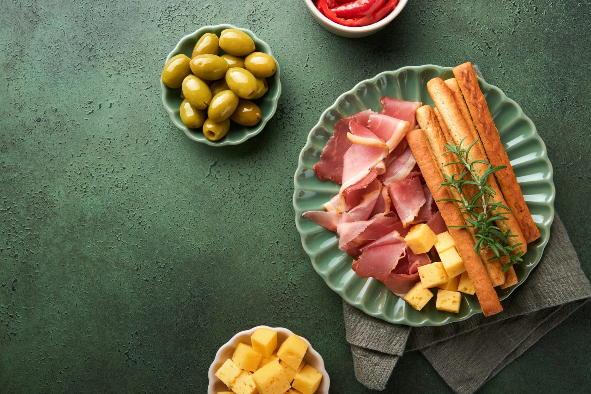 Charcuterie board with prosciutto, cheese cubes, breadsticks, and green olives on a textured green surface.