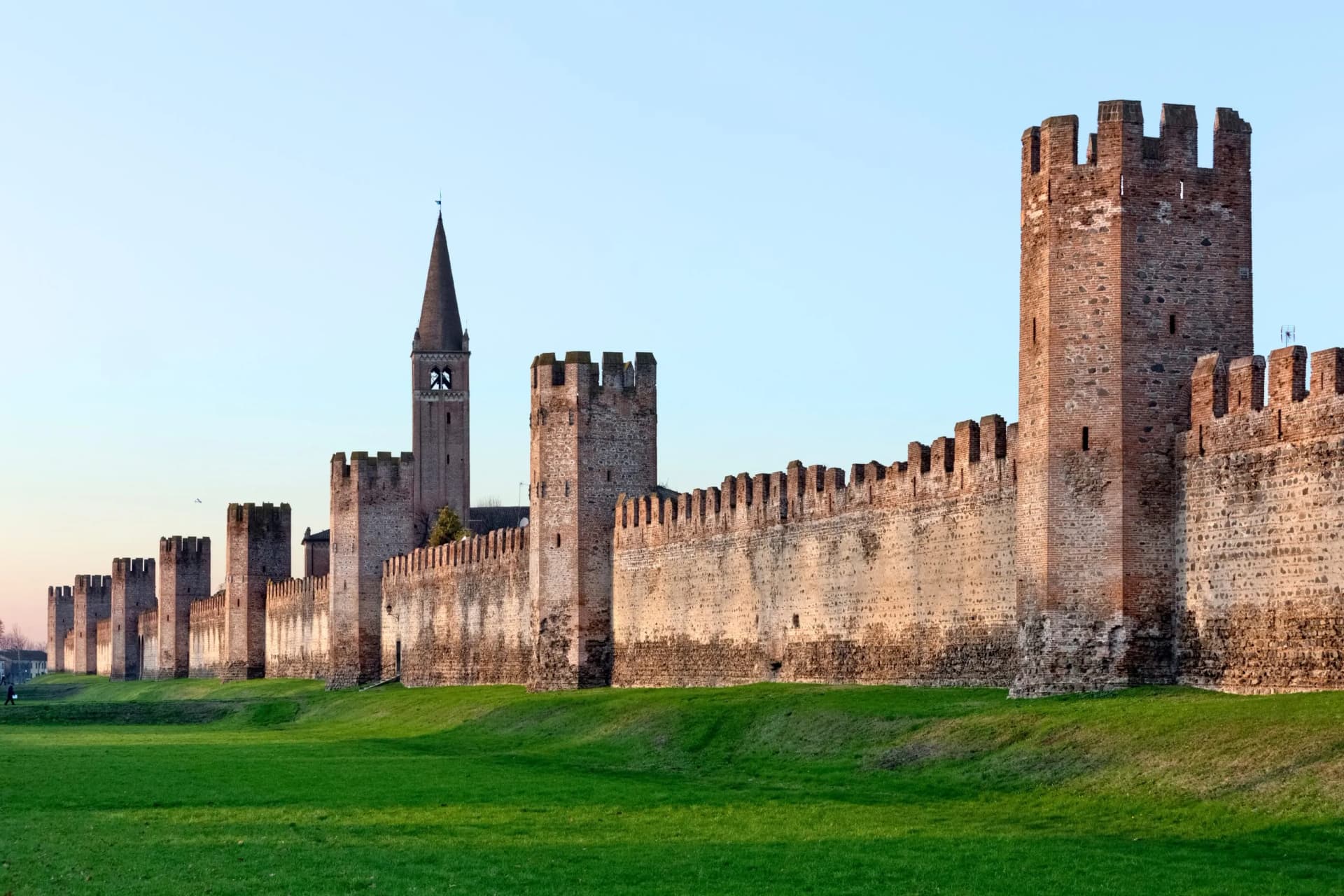 Medieval defensive walls and tall brick bell tower above a bright green grassy moat in Montagnana.