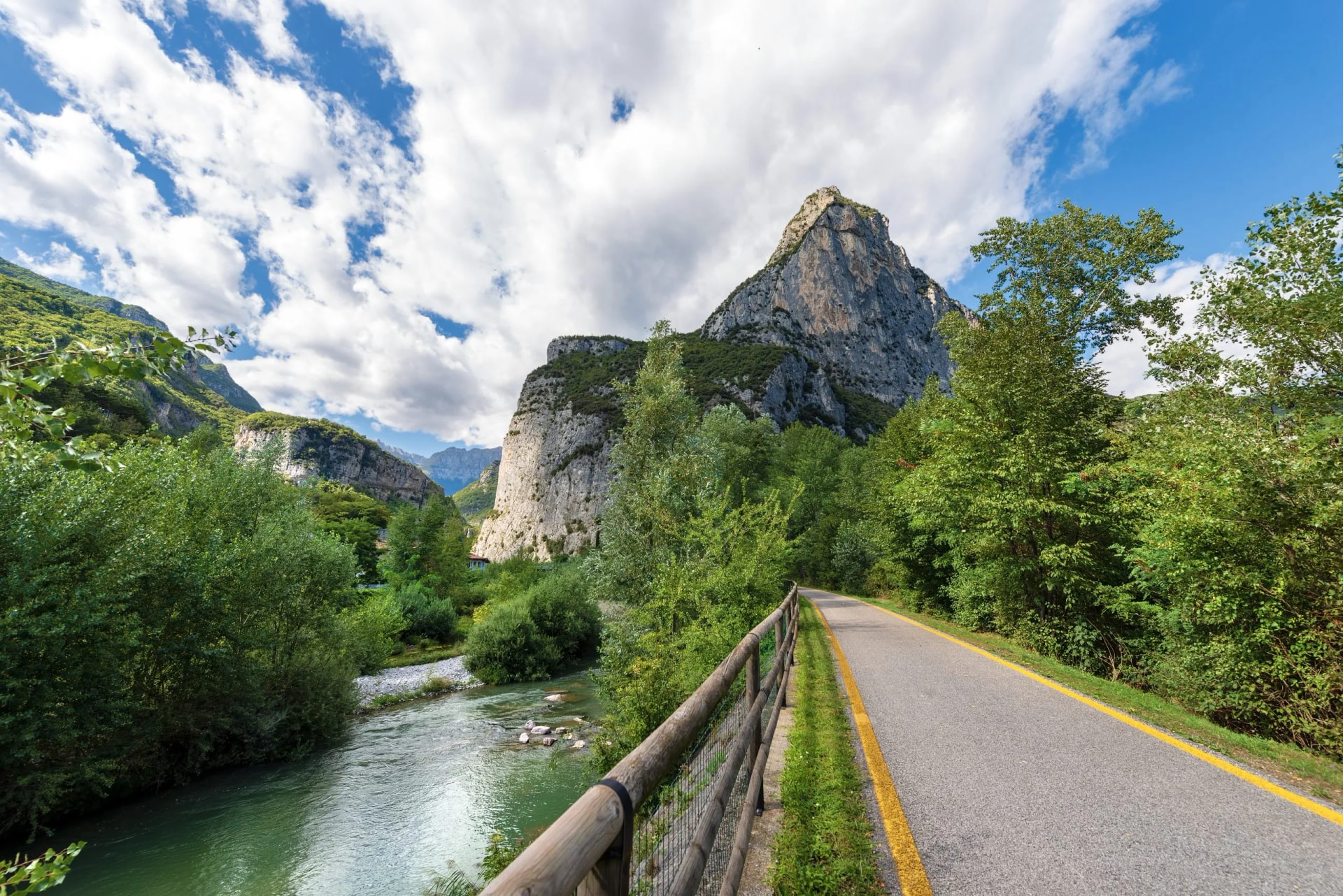 Cycling path by river next to sheer cliffs and green mountains under a cloudy blue sky.