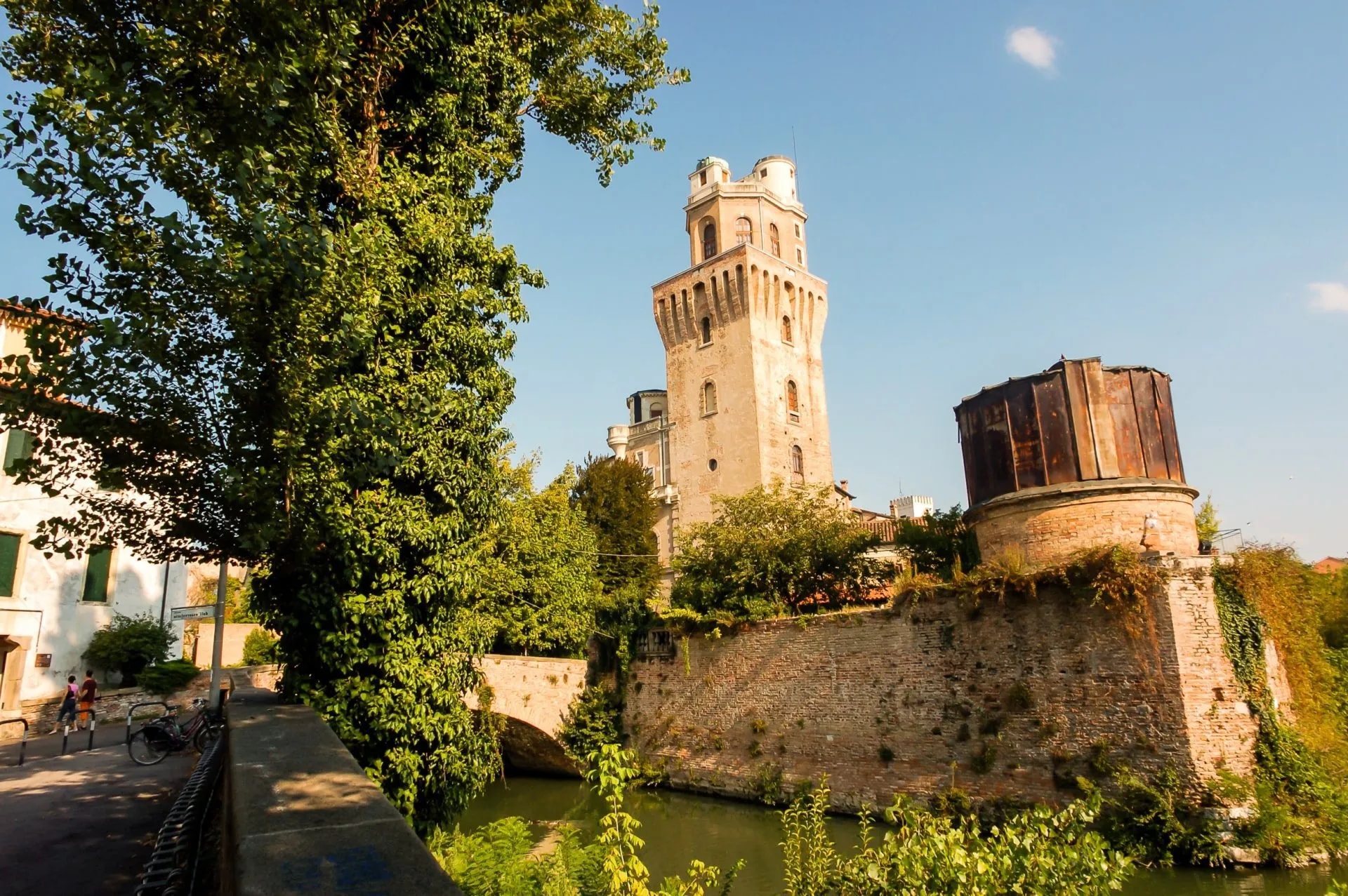 Tower and brick fortress wall next to canal in Padova with lush green trees.