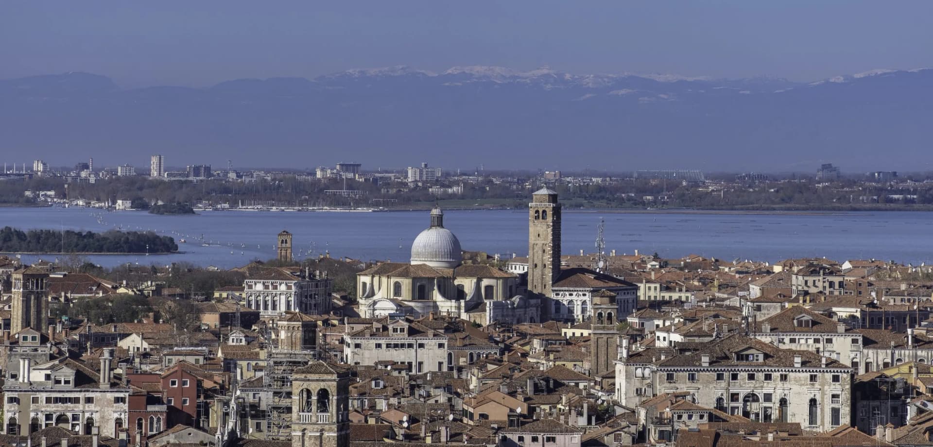 Rooftops of Venice with a large dome and bell tower, lagoon, and snowy mountains.