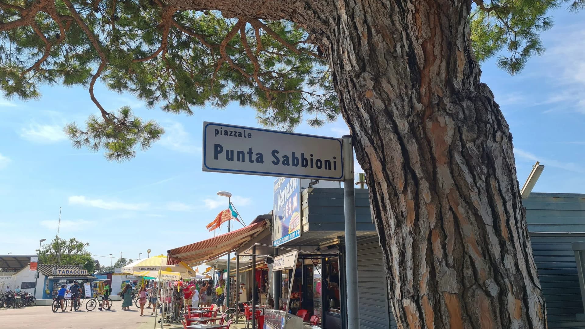 Piazzale Punta Sabbioni street sign next to large tree trunk with shops and cyclists nearby.