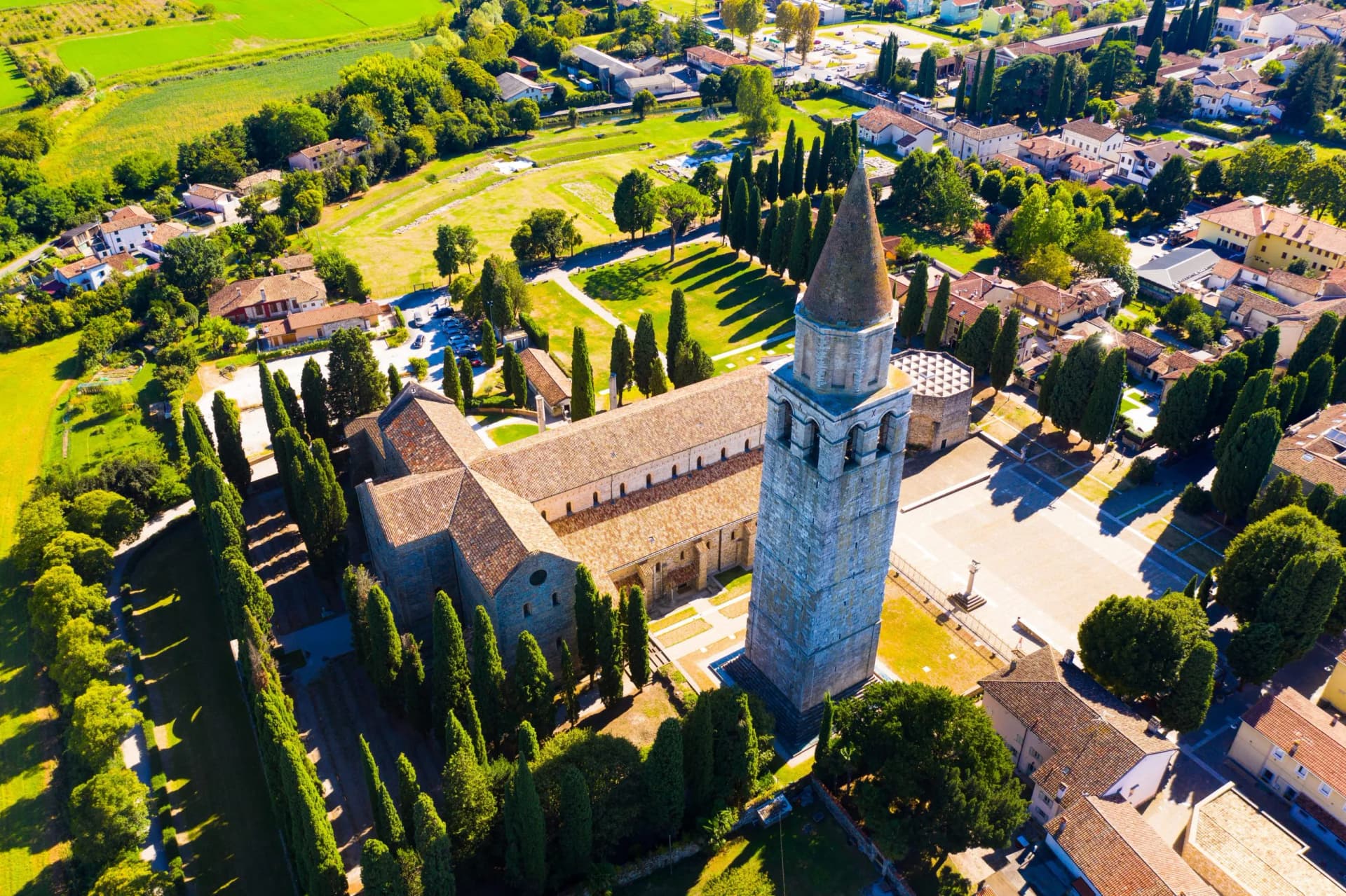 Aerial view of Aquileia Basilica and bell tower surrounded by cypress trees and green fields.