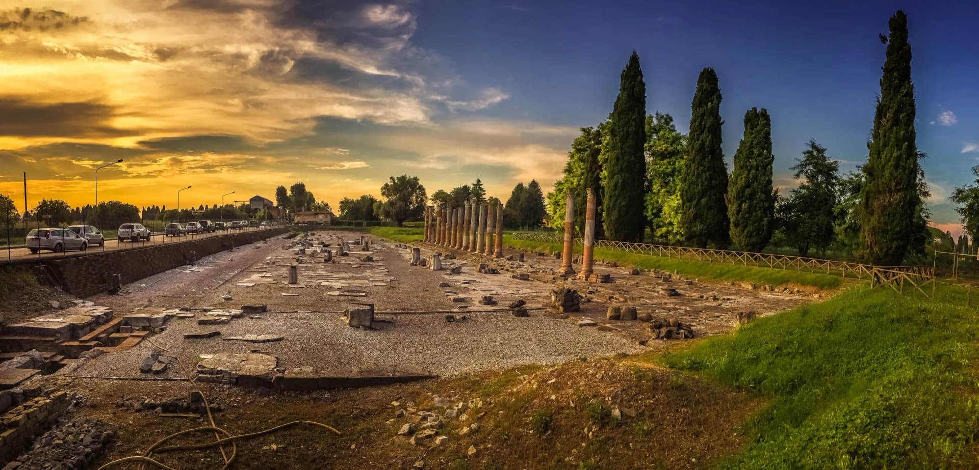 Roman ruins with standing columns, cypress trees, and traffic on a nearby road at sunset.
