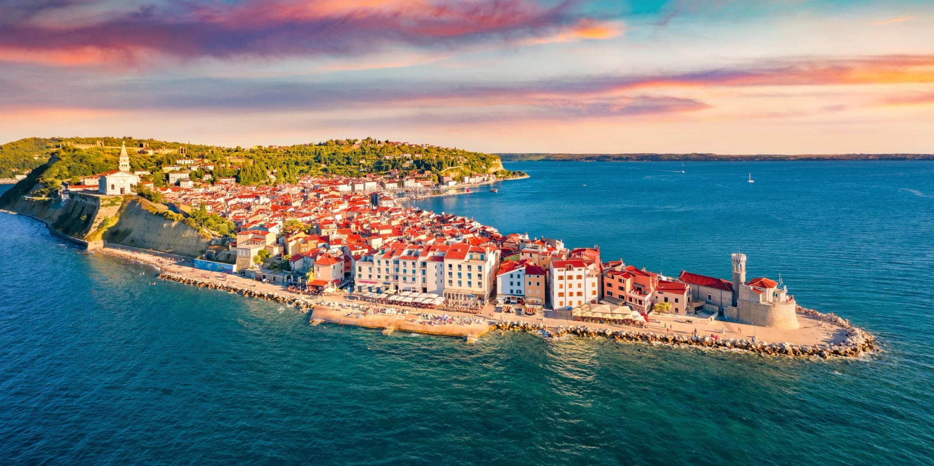 Coastal town with red roofs on a peninsula, set against blue sea and colorful sunset sky.