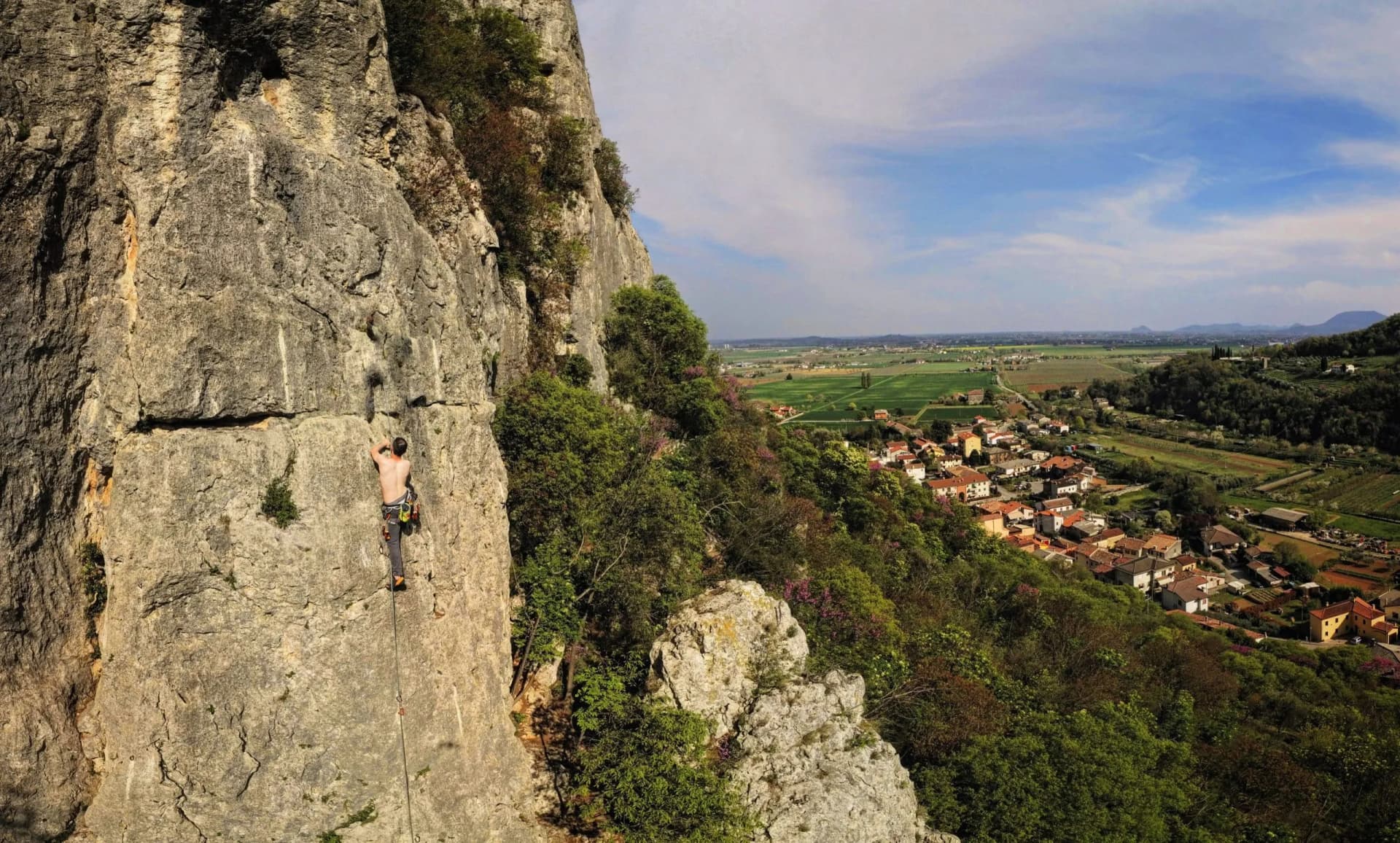 Rock climber ascending cliff face overlooking Lumignano village and green valley.