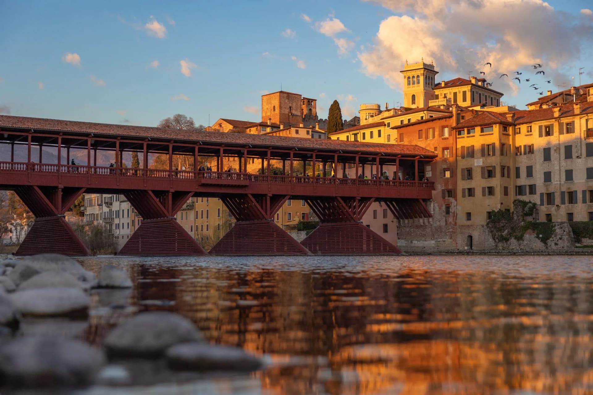 Covered wooden bridge over water with historic buildings at sunset in Bassano.