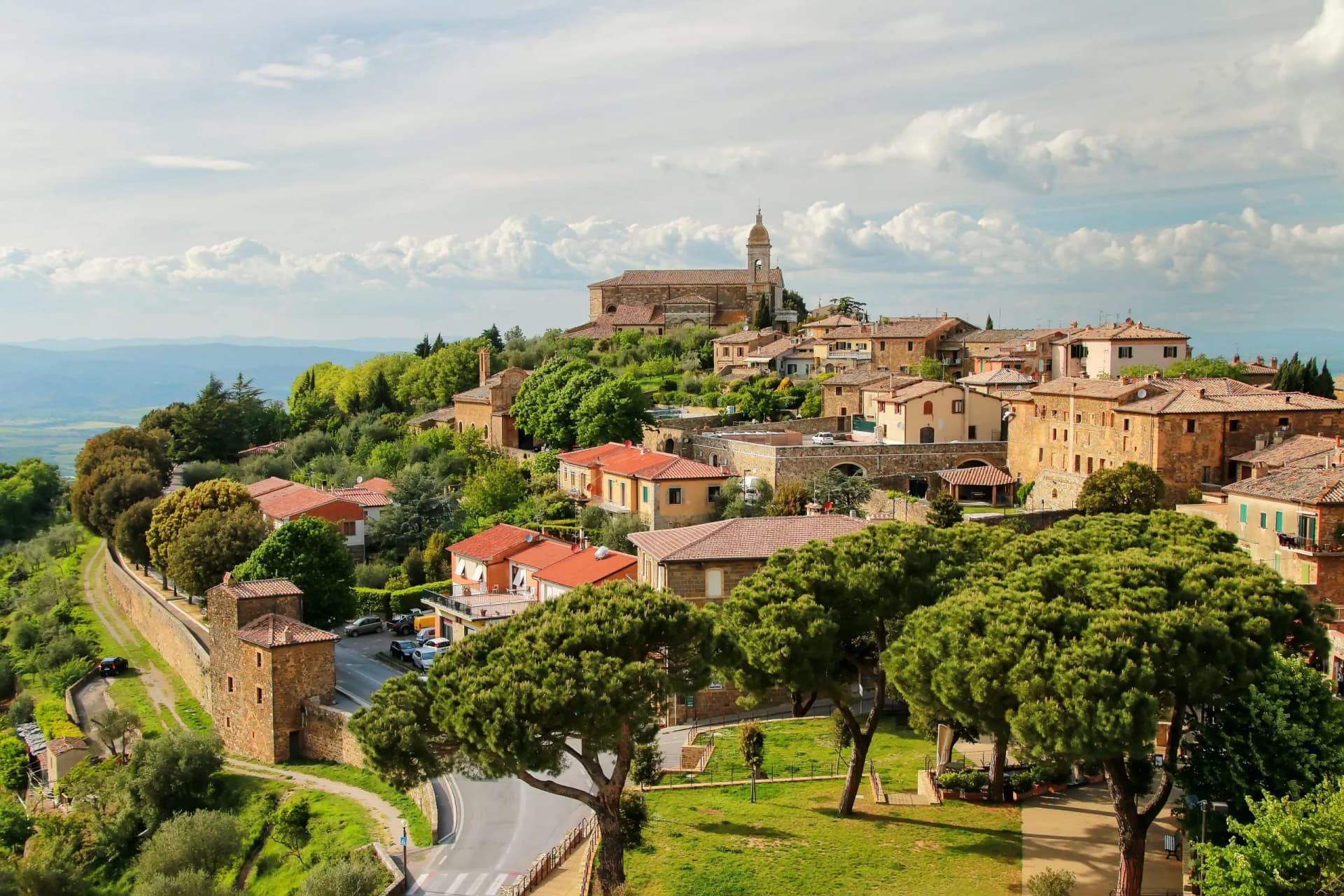Hilltop town with terracotta roofs, church spire, and green trees under a cloudy sky in Montalcino.